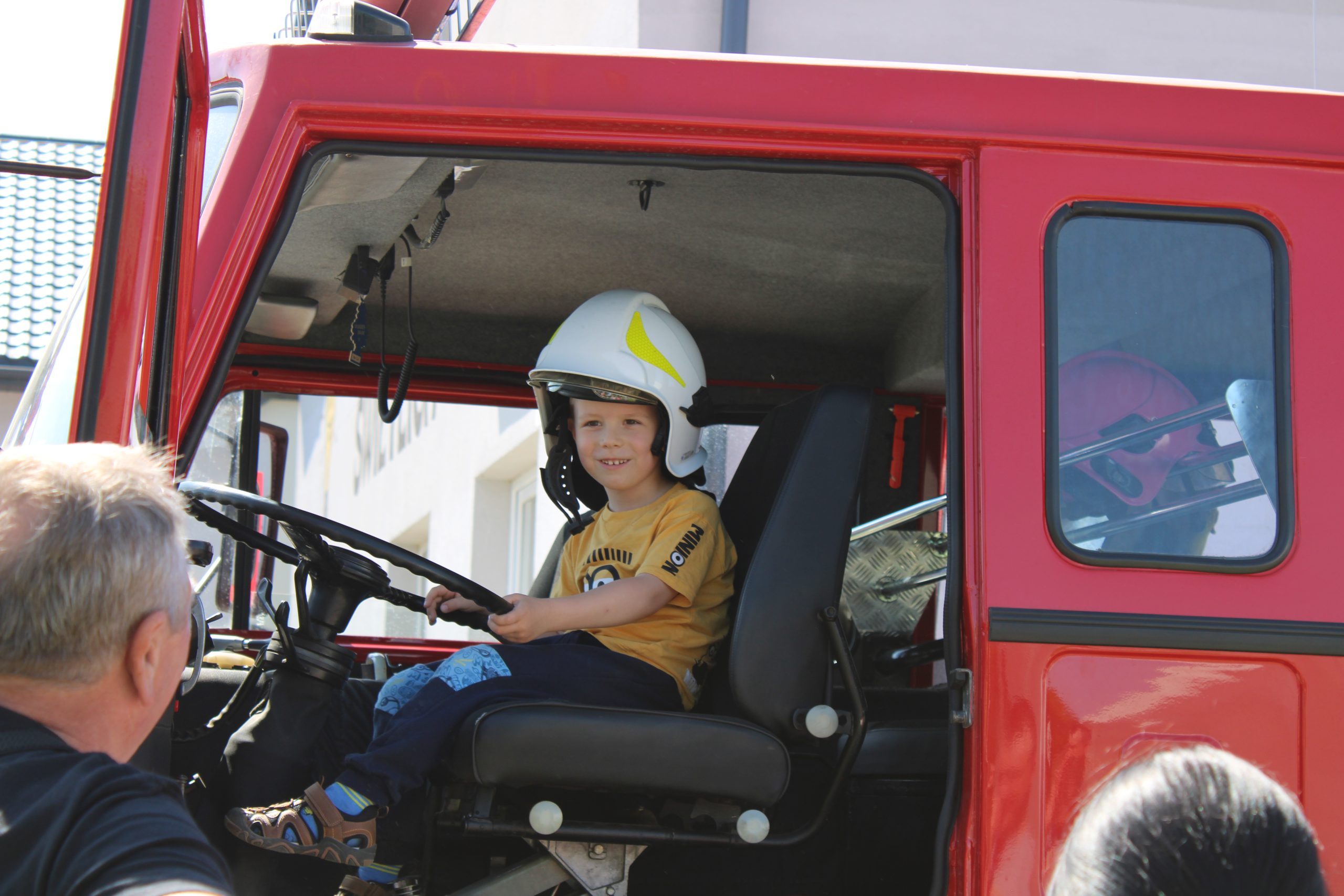 Na zdjęciu widoczny jest chłopiec w kasku siedzący w wozie strażackim, a przy nim stoi pan.