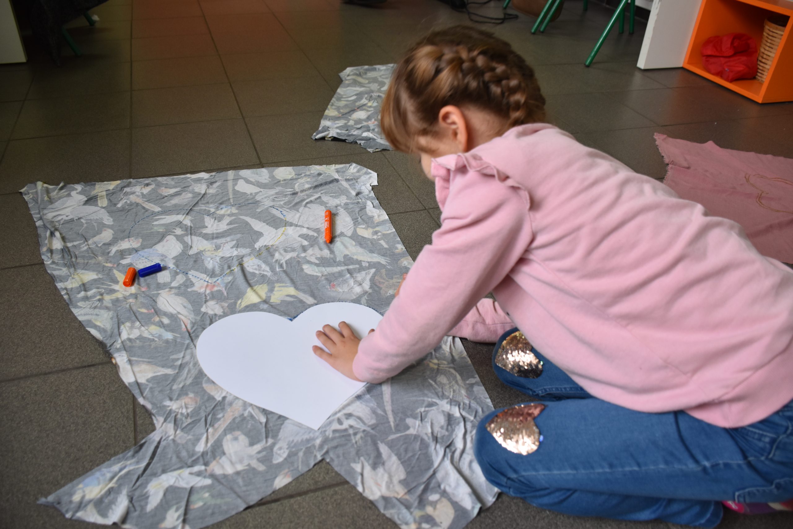 Na zdjęciu jest dziewczynka w wieku siedmiu lat o brązowych włosach i uplecionym z nich warkoczu. Ubrana jest w różową bluzkę i jeansowe spodnie z błyszczącymi sercami na kolanach. Przed nią na podłodze rozłożony jest materiał z wzorem w ptaki. Do materiału dziewczynka przykłada szablon w kształcie serca i odrysowuje kształt.