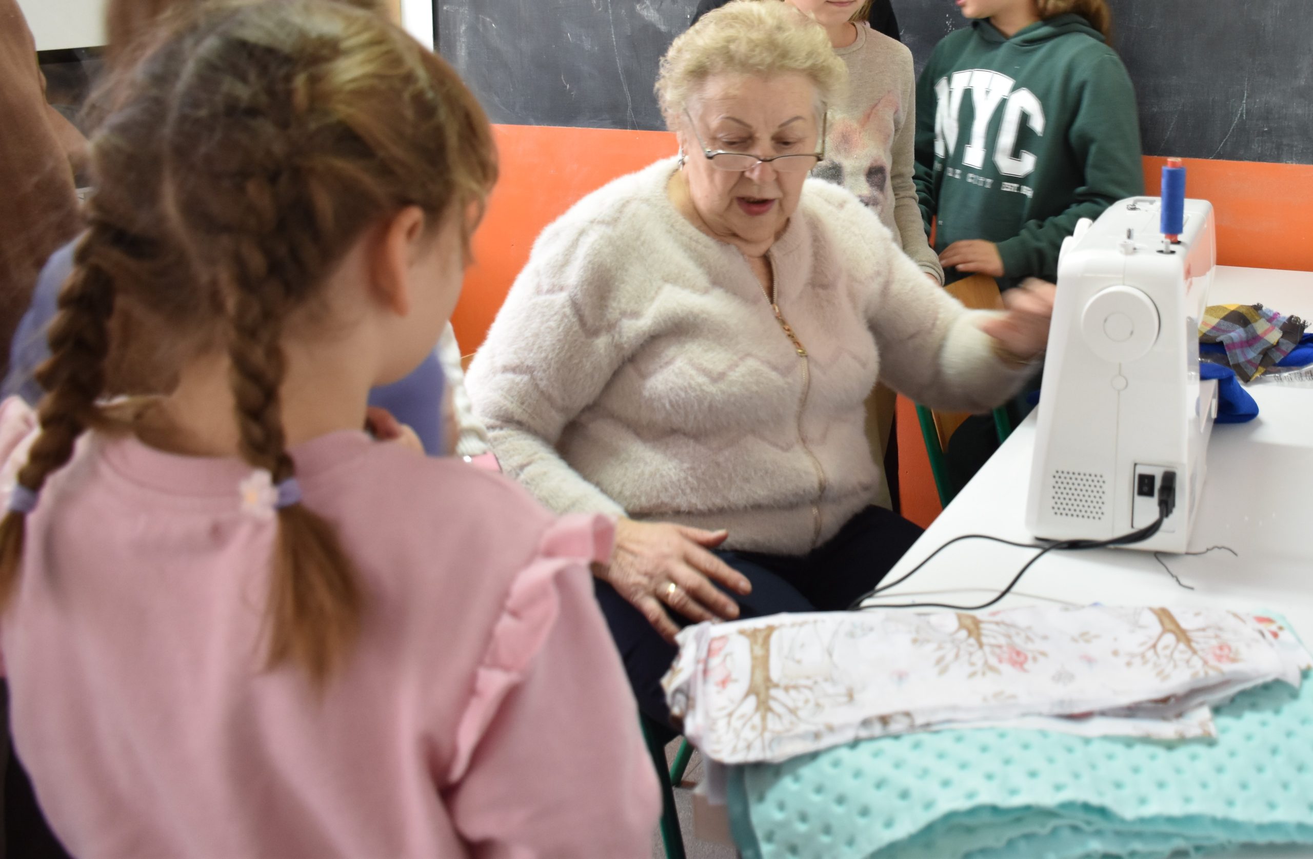 Na zdjęciu widać starszą Panią zwracającą się do grupy dziewczynek stojących wokół niej. Seniorka siedzi przy maszynie. Obok po prawej stronie leżą materiały. Starsza Pani ubrana jest w zasuwany sweterek o miłej strukturze i jasnym odcieniu. Ma założone okulary. Dziewczynka po lewej stronie ubrana jest w różową bluzę, zrobione warkoczyki i stoi przodem do Seniorki.