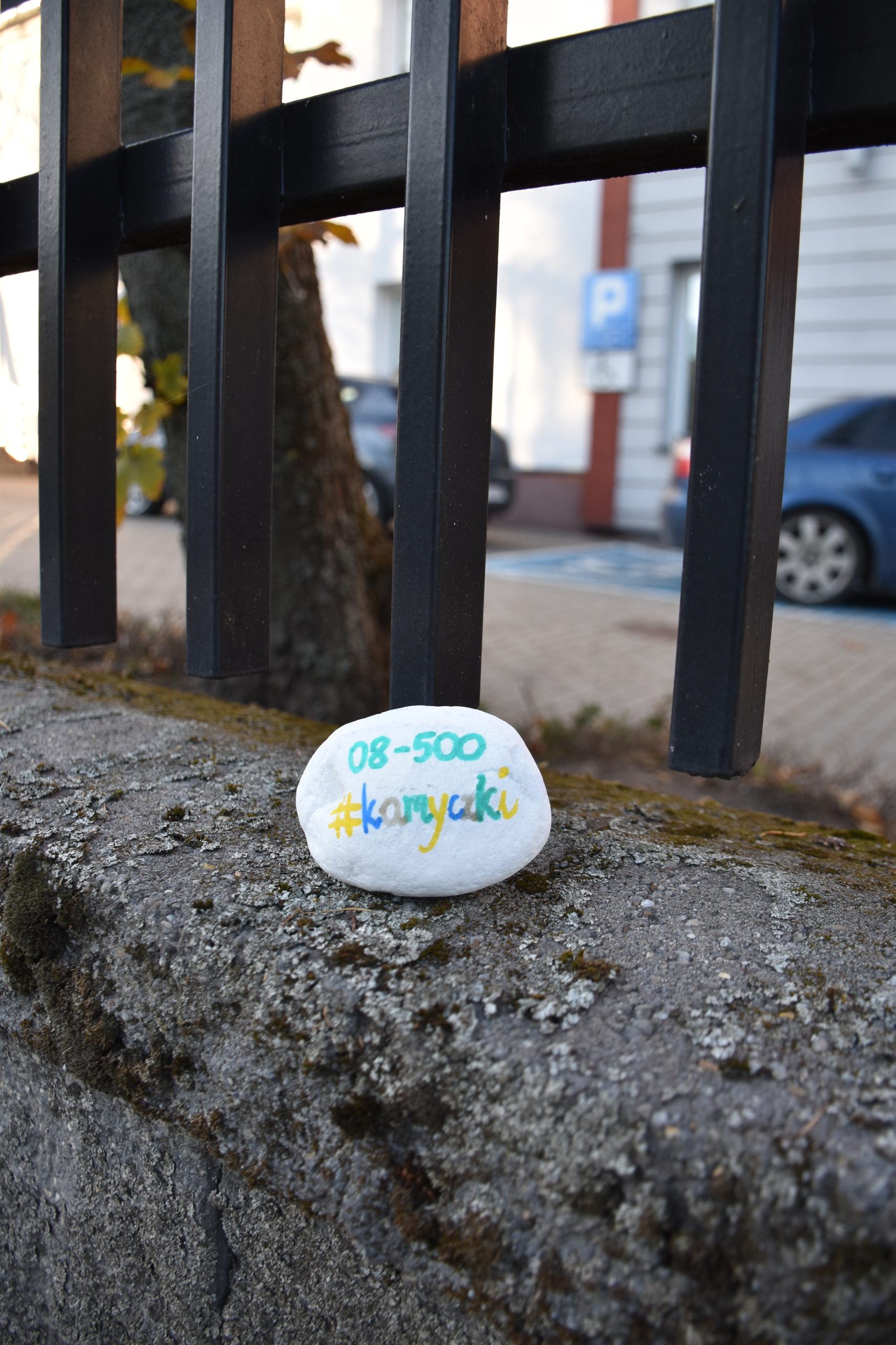 Na zdjęciu widać biały kamyk zawierający kolorowy napis 08-500 oraz #kamyczki. Kamyk leży na wysokim krawężniku tuż przy czarnym metalowym ogrodzeniu. Za ogrodzeniem widać drzewo, dwa samochody, budynek oraz znak określający miejsca parkingowe.