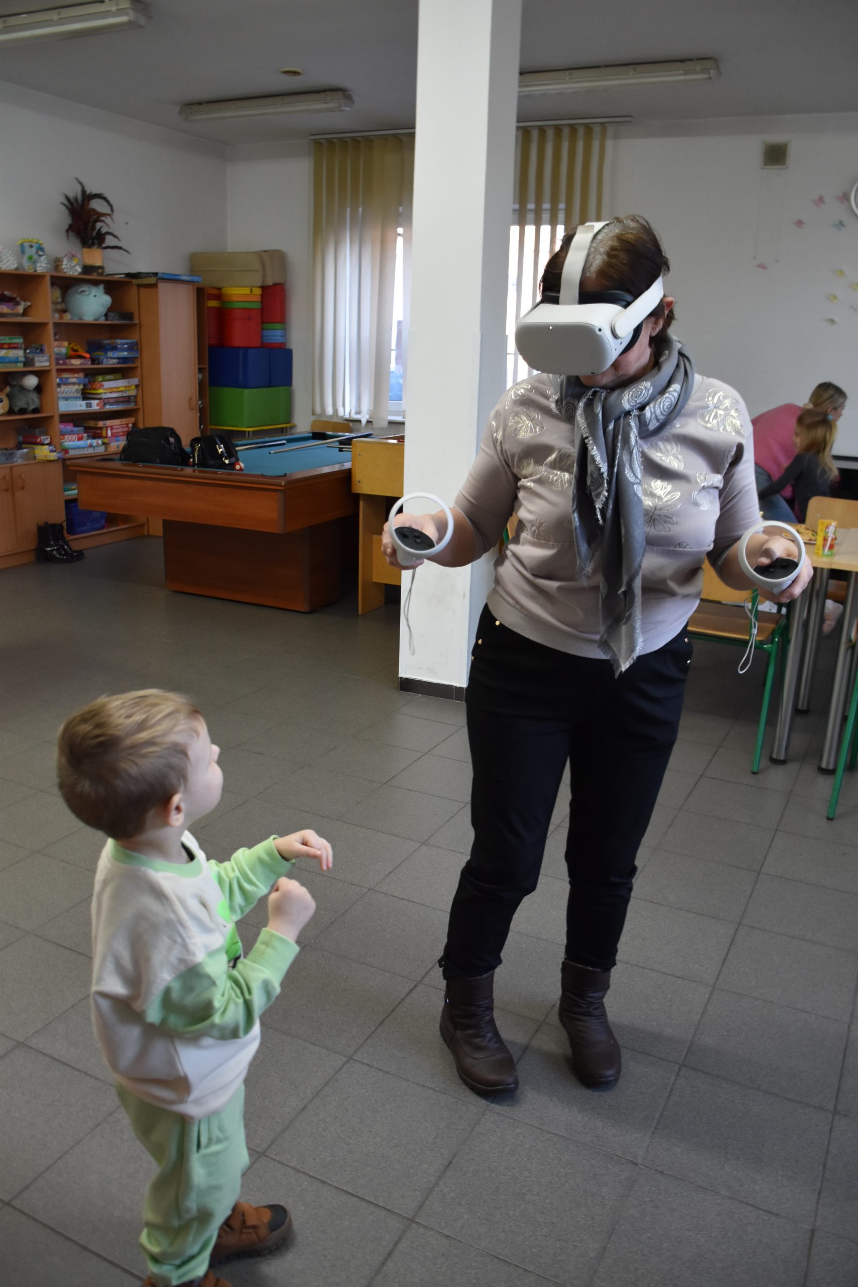 Na zdjęciu widać starszą kobietę w okularach VR. Kobieta ma ciemne spodnie i jasną bluzkę. Na szyi ma apaszkę. Obok niej stoi mały chłopiec, ma około dwóch lat. W tle widać regały z zabawkami, duże klocki, stół do bilarda, krzesełka i słup na środku sali. W tle jest też okno z roletą.