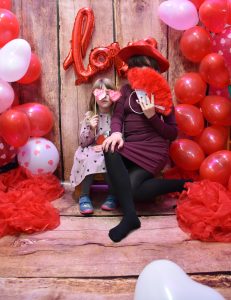 Na zdjęciu widać dwie dziewczynki robiące sobie zdjęcie na specjalnym tle. Tło jest koloru drewna. Za nimi widać balon "love". Dziewczynki siedzą na drewnianej ławeczce. Po bokach znajduje się duża ilość balonów czerwonych i kilka białych. Starsza dziewczynka po prawej stronie ma bordową sukienkę i ciemne rajstopy. Ma na głowie czerwony kapelusz, a twarz zakrywa piórkowym czerwonym wachlarzem. Młodsza dziewczynka w sukience w czarne serduszka trzyma okulary serduszka na patyczku.