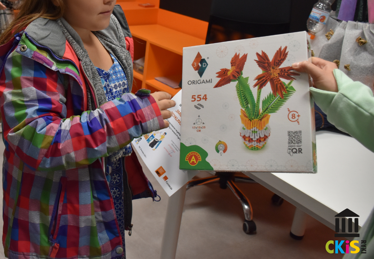 Na zdjęciu widać dziecko ubrane w kolorową kurtkę w kratę (czerwone, niebieskie, zielone i szare kwadraty), które trzyma pudełko z zestawem do origami 3D. Pudełko jest białe, a na nim widoczny jest gotowy model – pomarańczowe kwiaty z zielonymi liśćmi w kolorowej, żółto-białej doniczce. Na opakowaniu jest napis „ORIGAMI 3D”, numer „554”, a także oznaczenie, że zestaw zawiera elementy do składania i jest przeznaczony dla dzieci od 8 lat wzwyż. Z boku pudełka znajduje się kod QR. Zdjęcie zostało wykonane w pomieszczeniu – w tle widać fragment białego stołu. W prawym dolnym rogu widnieje kolorowe logo z napisem „CKiS Ryki”.