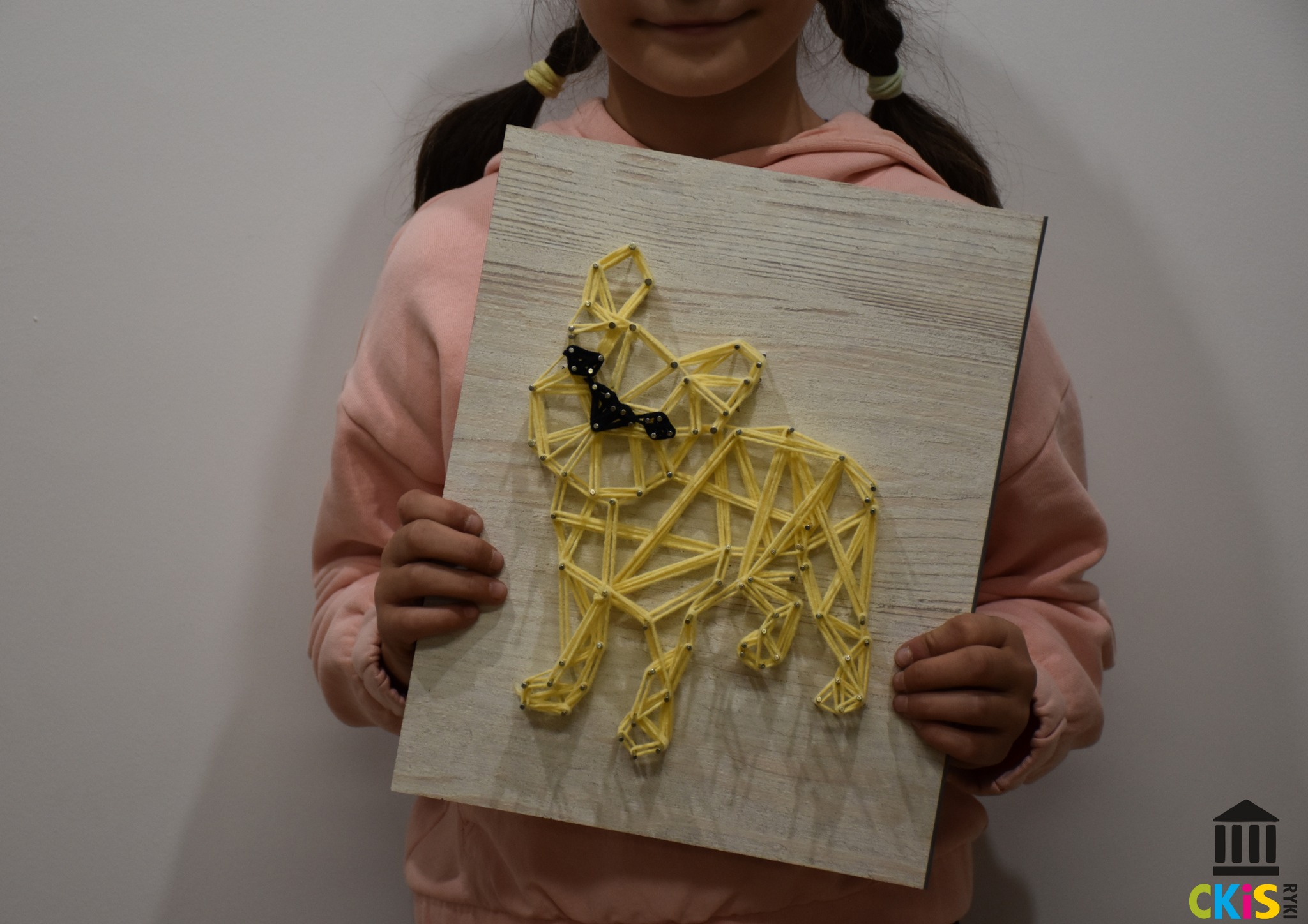 Na zdjęciu widać dziecko, które trzyma obrazek wykonany metodą string-art . Widać na nim pieska wykonanego z żółtych nici.