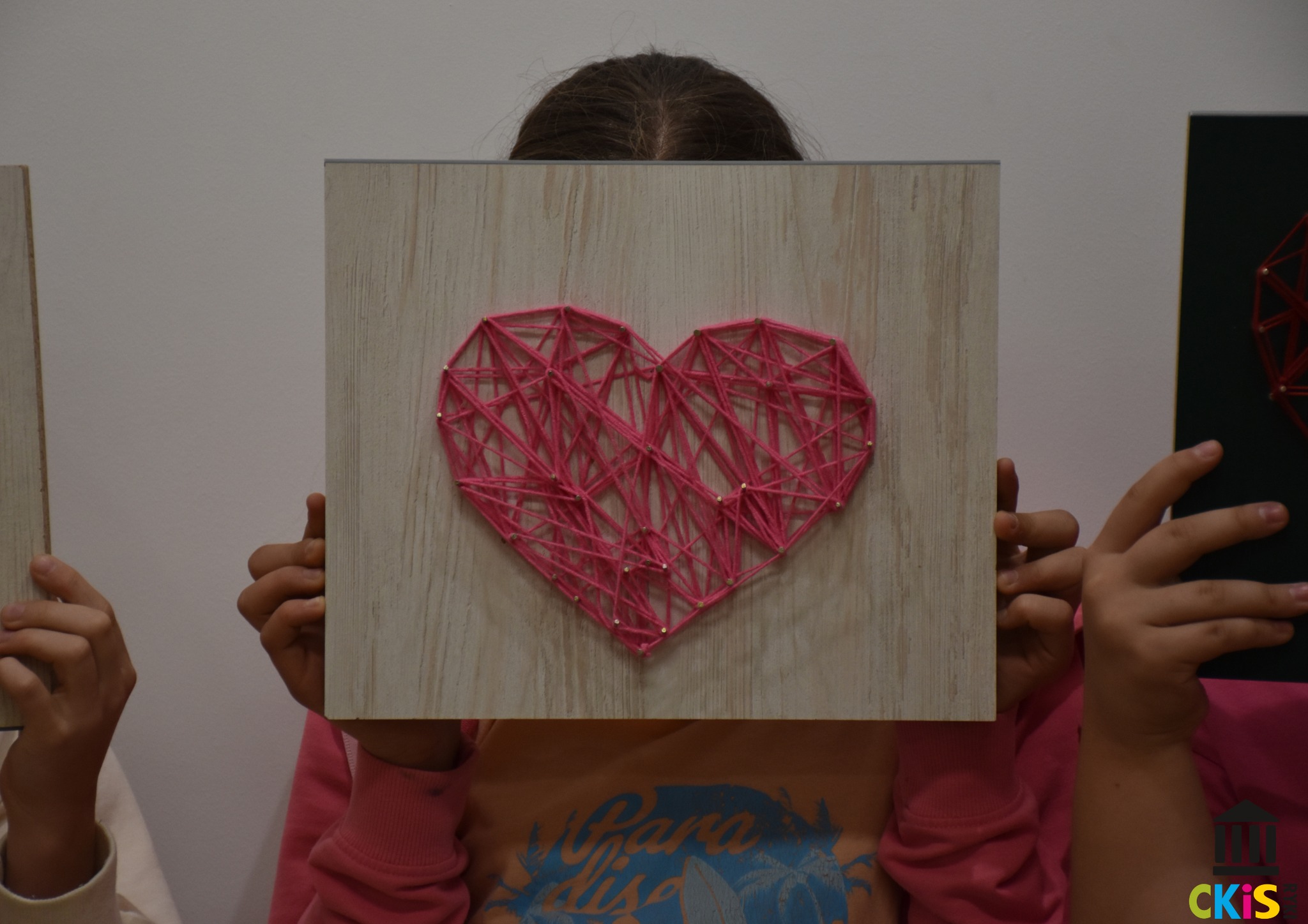Na zdjęciu widać dziecko, które trzyma obrazek wykonany metodą string-art. Na obrazku jest serce, wykonane z różowych nici.