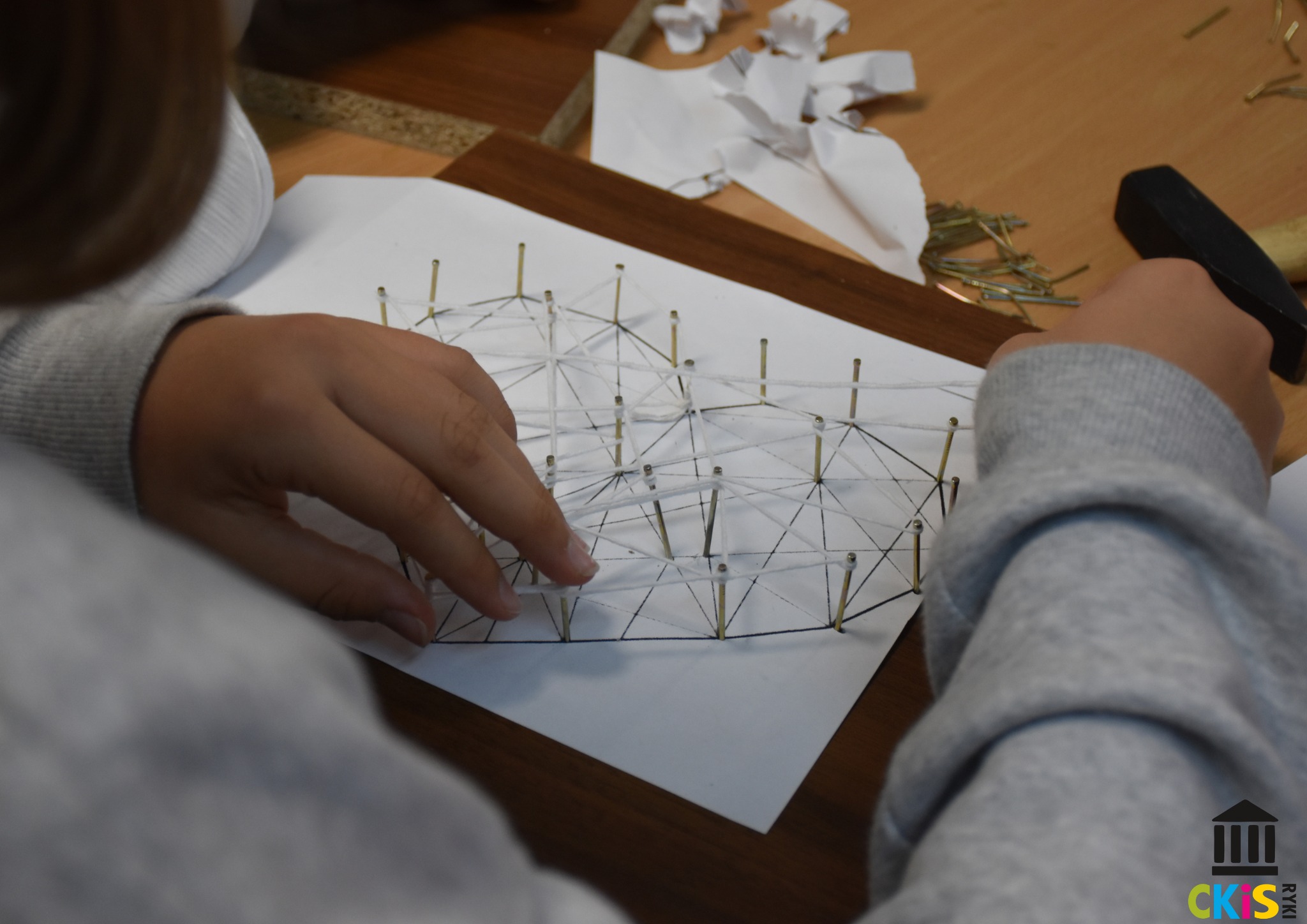 Na zdjęciu widzimy zbliżenie na dłonie dziecka pracującego nad projektem typu string art – czyli sztuką tworzenia obrazów za pomocą nici rozciąganych pomiędzy gwoździami wbitymi w drewnianą tablicę. Jedną ręką przytrzymuje nici które są już rozciągnięte między gwoździami, tworząc skomplikowany geometryczny wzór.Gwoździe są wbite w tablicę na podstawie papierowego szablonu.