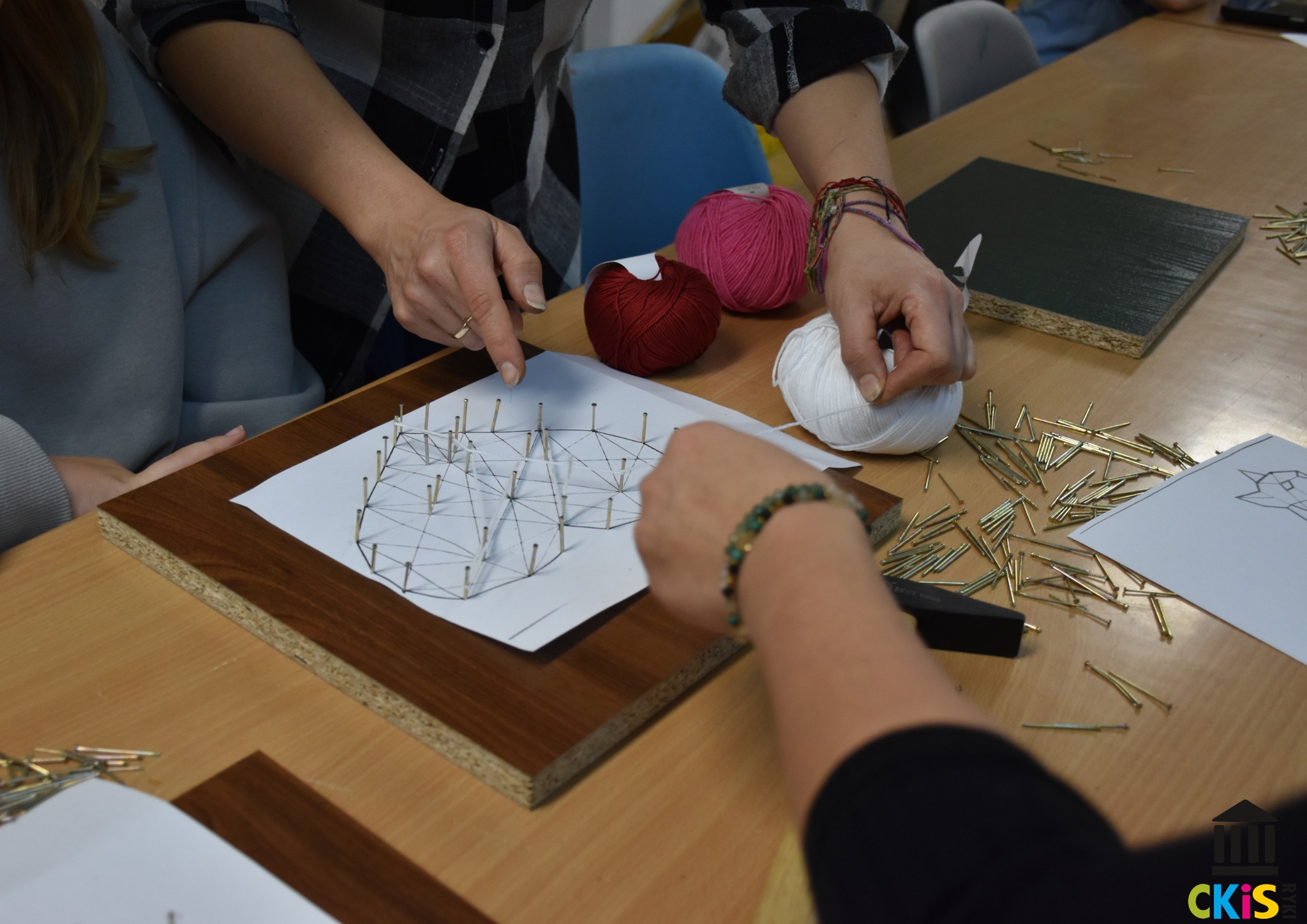 Na zdjęciu widzimy grupę osób uczestniczących w warsztatach rękodzielniczych. Skupiają się one na tworzeniu obrazu techniką string art, czyli sztuki tworzenia wzorów przy użyciu nici rozciągniętych między gwoździami wbitymi w deskę. Na stole leży prostokątna płyta wiórowa z ramką z ciemniejszego drewna, do której przymocowany jest biały arkusz papieru z konturami wzoru serca. W kontury zostały wbite krótkie, złote gwoździe. Jedna z osób trzyma białą włóczkę, a druga wskazuje palcem na wzór, tłumacząc coś. Obok leżą kłębki włóczki w kolorach białym, czerwonym i różowym oraz duża ilość niewbitych gwoździ.