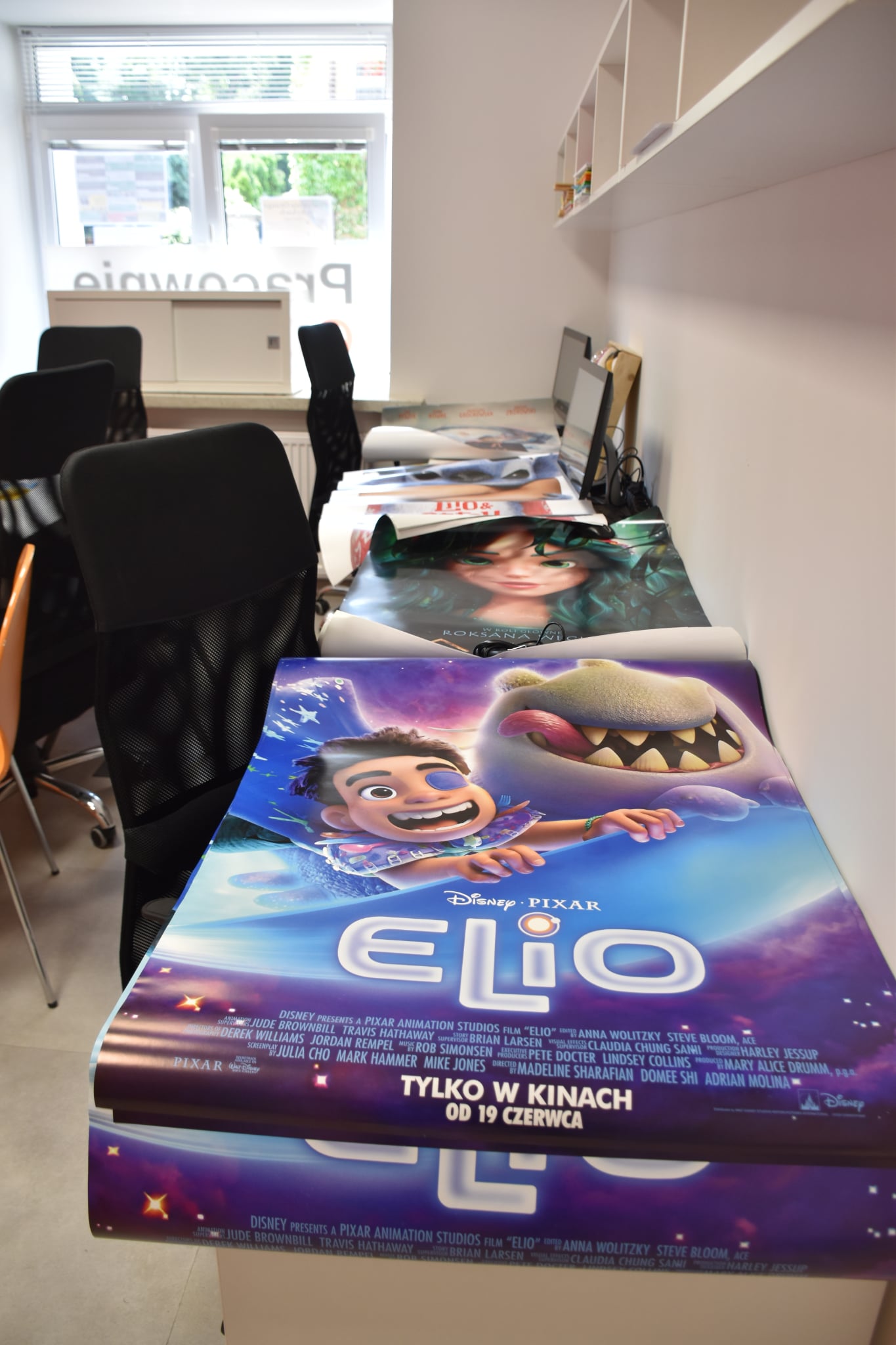 Na biurku w sali ułożono w stosie duże filmowe plakaty. Najbliżej widoczny jest plakat z filmu animowanego „Elio” w intensywnych kolorach niebieskiego i fioletowego. To właśnie z takich plakatów dzieci wycinały materiały do wykonania swoich wiatraków.