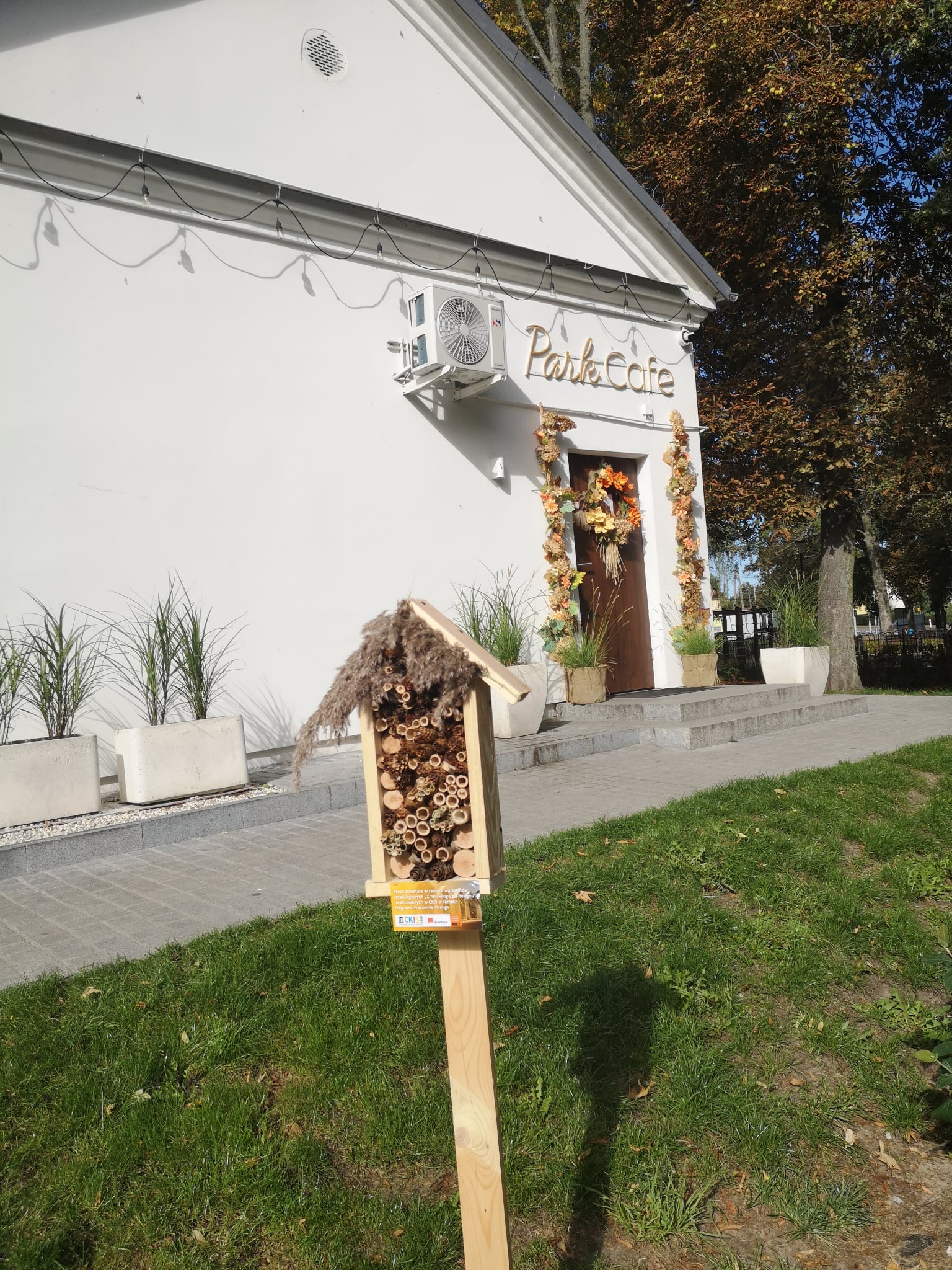 Na zdjęciu widzimy teren zielony przy białym budynku z napisem „Park Cafe”. Na pierwszym planie stoi domek dla owadów zapylających, zamocowany na drewnianym słupku wbitym w trawnik. Domek ma kształt pionowego prostokąta z daszkiem i jest wypełniony naturalnymi materiałami: kawałkami drewna, bambusowymi rurkami, szyszkami i suchymi roślinami.Na dole domku przymocowana jest mała tabliczka informacyjna.