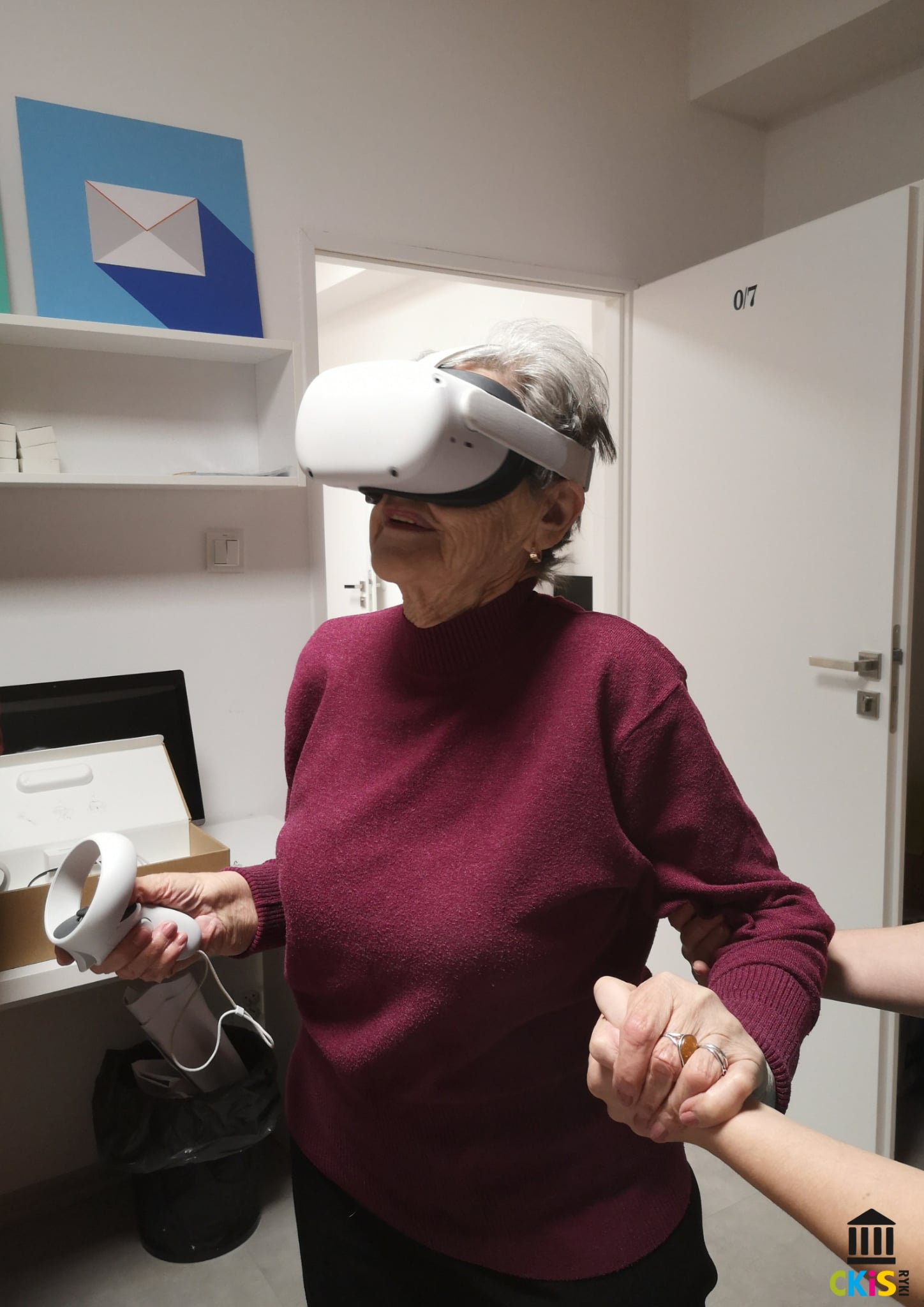 Na zdjęciu widać starszą kobietę korzystającą z zestawu wirtualnej rzeczywistości. Ma na głowie duże, białe gogle VR, a w jednej ręce trzyma kontroler. Ubrana jest w bordowy sweter. Obok niej znajduje się druga osoba, która trzyma ją za ramię i za dłoń — prawdopodobnie po to, by zapewnić jej wsparcie i bezpieczeństwo podczas korzystania z urządzenia.