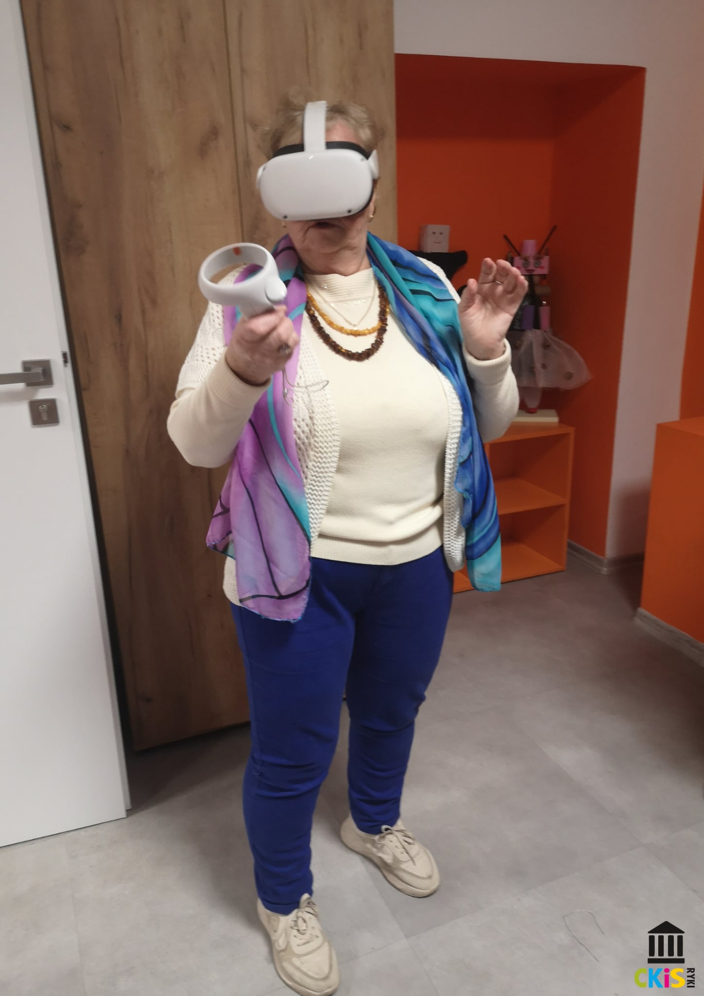 Na zdjęciu widać starszą kobietę korzystającą z gogli wirtualnej rzeczywistości. Ma na głowie duże, białe gogle VR, a w jednej ręce trzyma kontroler, którym najwyraźniej wykonuje ruch lub bierze udział w jakiejś interakcji. Drugą rękę ma uniesioną w lekkim geście, jakby reagowała na coś, co widzi w wirtualnym świecie.