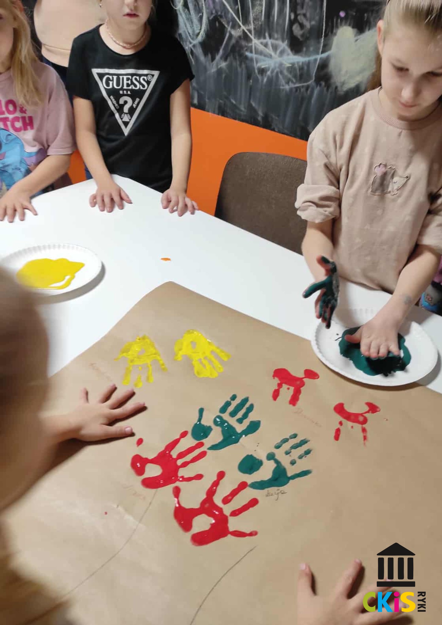 na zdjęciu widać dzieci, które maczają dłonie w farbie, obok nich leży obraz z odciśniętymi dłońmi na papierze