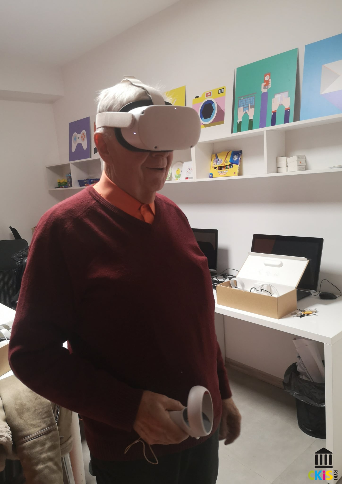 Na zdjęciu widać starszego mężczyznę korzystającego z zestawu wirtualnej rzeczywistości. Ma na głowie białe gogle VR, a w jednej ręce trzyma kontroler. Wygląda na zaangażowanego i zaciekawionego tym, co widzi w wirtualnym świecie.