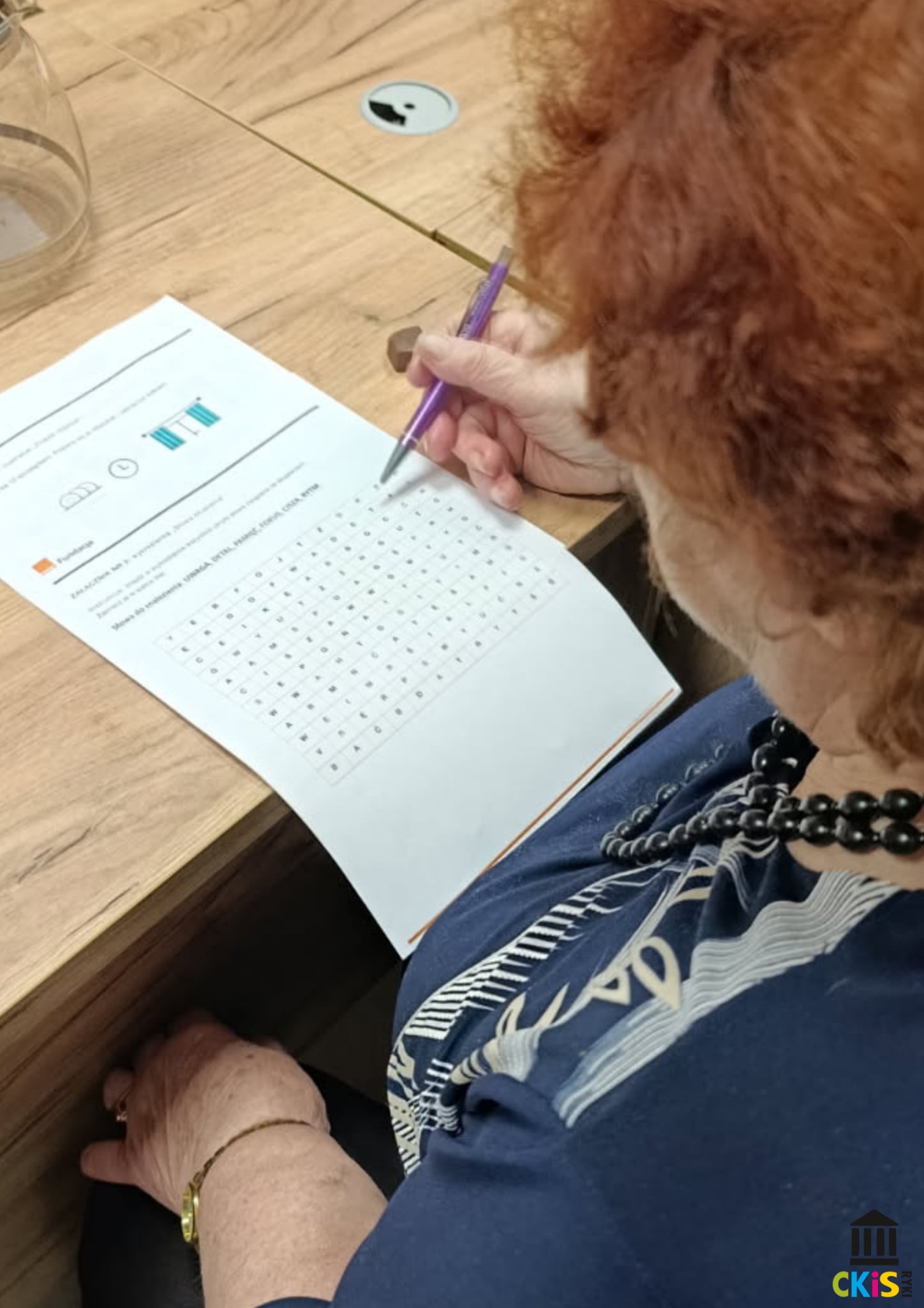 Na zdjęciu widać starszą kobietę siedzącą przy drewnianym stole i rozwiązującą zadanie na kartce. Trzyma fioletowy długopis i pochyla się nad arkuszem, który wygląda jak łamigłówka typu „word search” (wyszukiwanka słów). Kobieta ma rude, kręcone włosy i nosi granatową bluzkę oraz czarny naszyjnik z koralików.