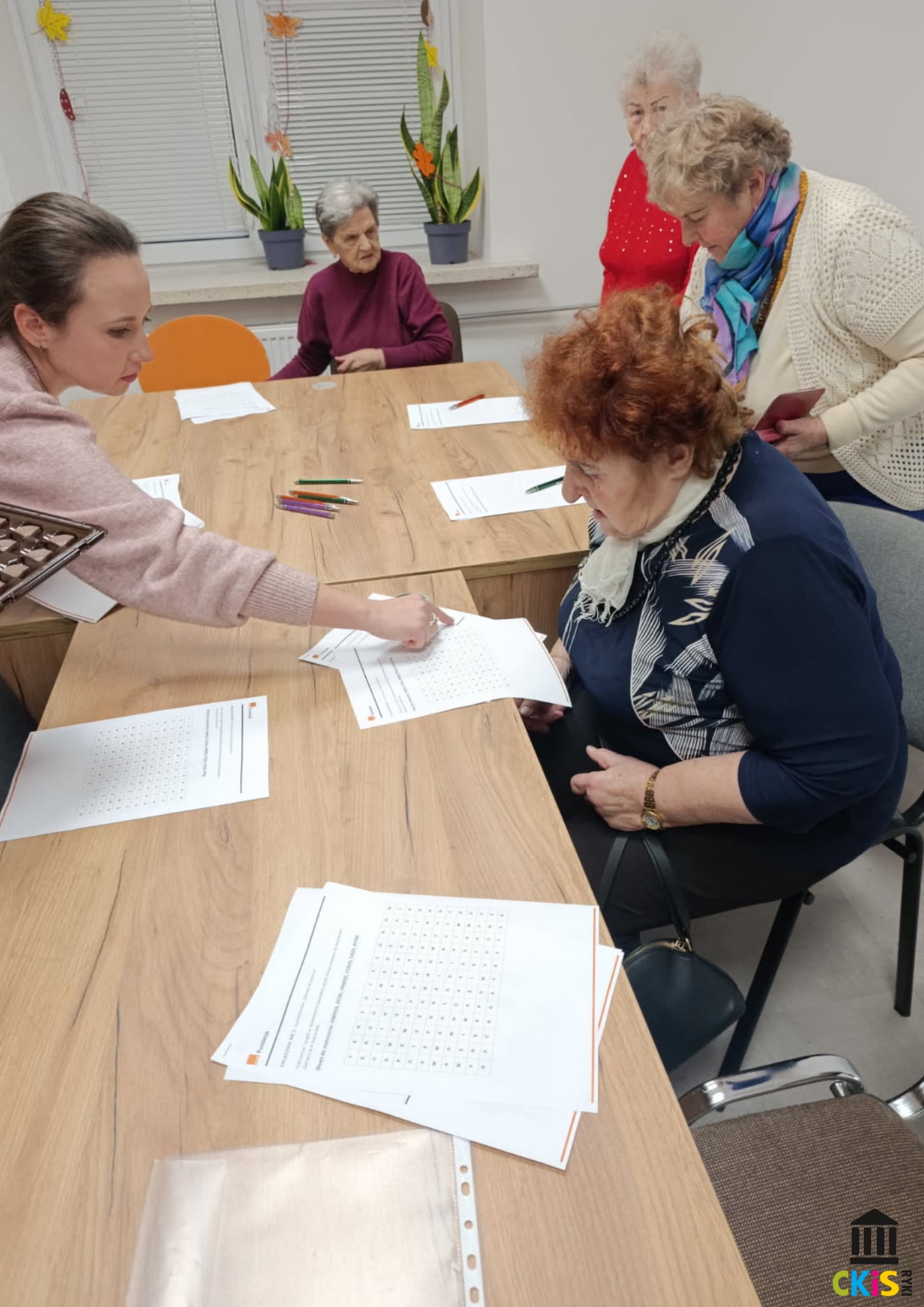 Na zdjęciu widać grupę starszych osób biorących udział w zajęciach przy stole. Siedzą i stoją wokół dużego drewnianego stołu, na którym leżą kartki z zadaniami – wyglądają jak łamigłówki i wykreślanki.