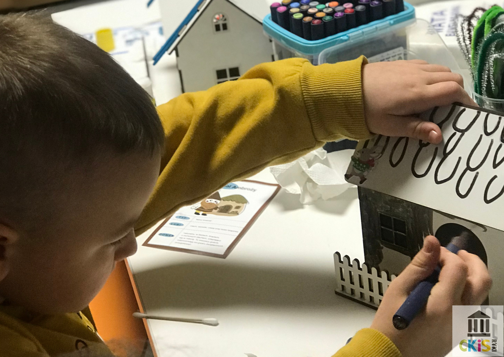 Na obrazku widać dziecko siedzące przy stole i skupione na pracy plastycznej. Dziecko ozdabia mały, drewniany domek, trzymając w jednej ręce marker lub flamaster, a drugą stabilizując domek. Ma na sobie żółtą bluzę. Domek ma biały dach z narysowanym wzorem oraz małe okna i drzwi. Na stole wokół znajdują się różne materiały plastyczne: flamastry w pojemniku, wyciory kreatywne, patyczki higieniczne oraz kartki z instrukcjami lub zadaniami. W tle widać także inne podobne domki, co sugeruje, że są to zajęcia grupowe lub warsztaty kreatywne. Cała scena pokazuje spokojną, twórczą atmosferę i koncentrację dziecka na wykonywanej pracy.