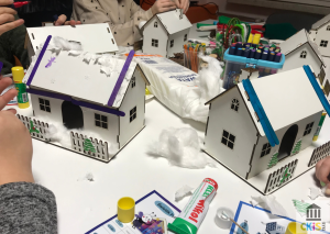 Na obrazku widać stół, przy którym dzieci wykonują prace plastyczne. Na stole stoją małe, białe, drewniane domki, które są ozdabiane jak zimowe lub świąteczne chatki. Dachy domków są przyklejone patyczkami w kolorach fioletowym i niebieskim, a na nich znajduje się watolina imitująca śnieg. Wokół domków leżą kawałki waty, klej w sztyfcie, flamastry, kredki, wyciory kreatywne oraz inne materiały plastyczne.