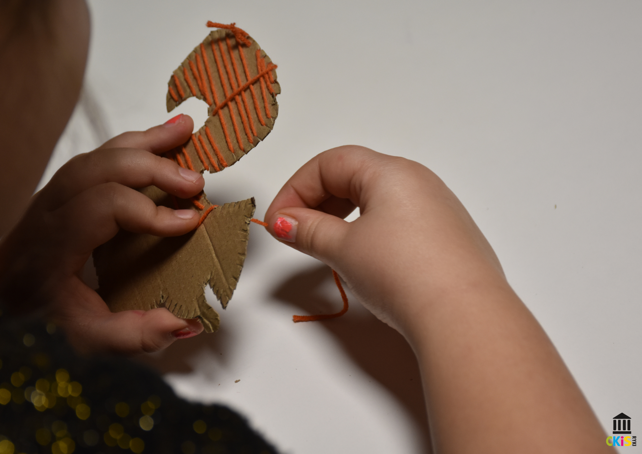 Na obrazku widać zbliżenie na dłonie dziecka wykonującego pracę plastyczną. Dziecko trzyma wycięte z tektury kształt wiewiórki. Przez otwory w tekturze przeciąga pomarańczową włóczkę, Tektura jest brązowa, a kontrastująca z nią włóczka ma intensywny, pomarańczowy kolor. Całość leży na jasnym, gładkim tle, co podkreśla skupienie na rękach i wykonywanej czynności.