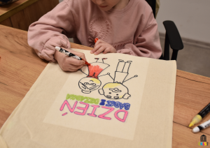 Na zdjęciu widać dziecko siedzące przy drewnianym stole, które z dużym skupieniem rysuje markerem na jasnej, materiałowej torbie.Na torbie znajduje się kolorowy, dziecięcy rysunek przedstawiający babcię i dziadka oraz napis „DZIEŃ BABCI I DZIADKA”.