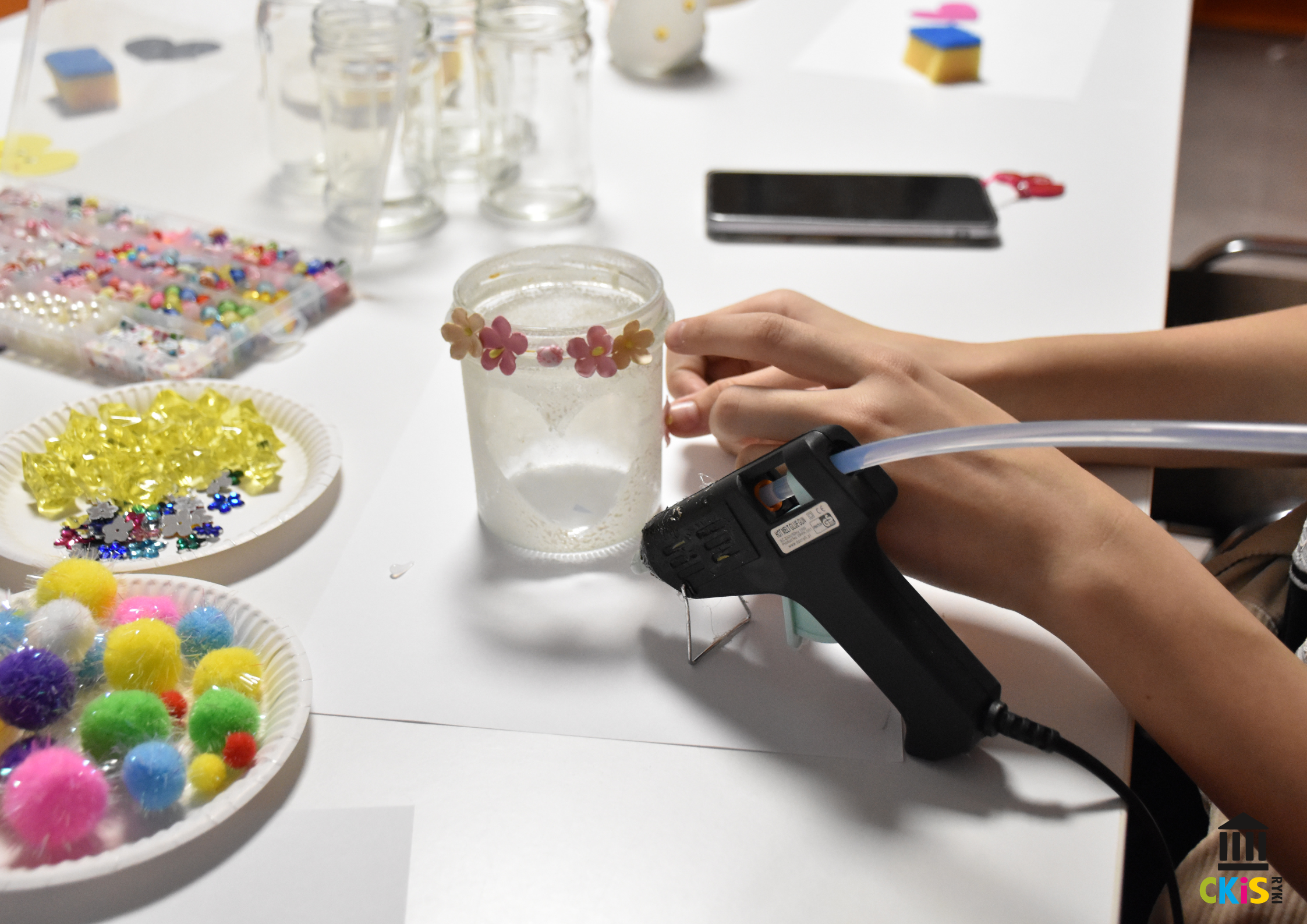 na zdjęciu widać słoik ozdobiony kwiatkami, dłoń dziewczynki trzyma pistolet do kleju na gorąco, przykleja kwiatuszki