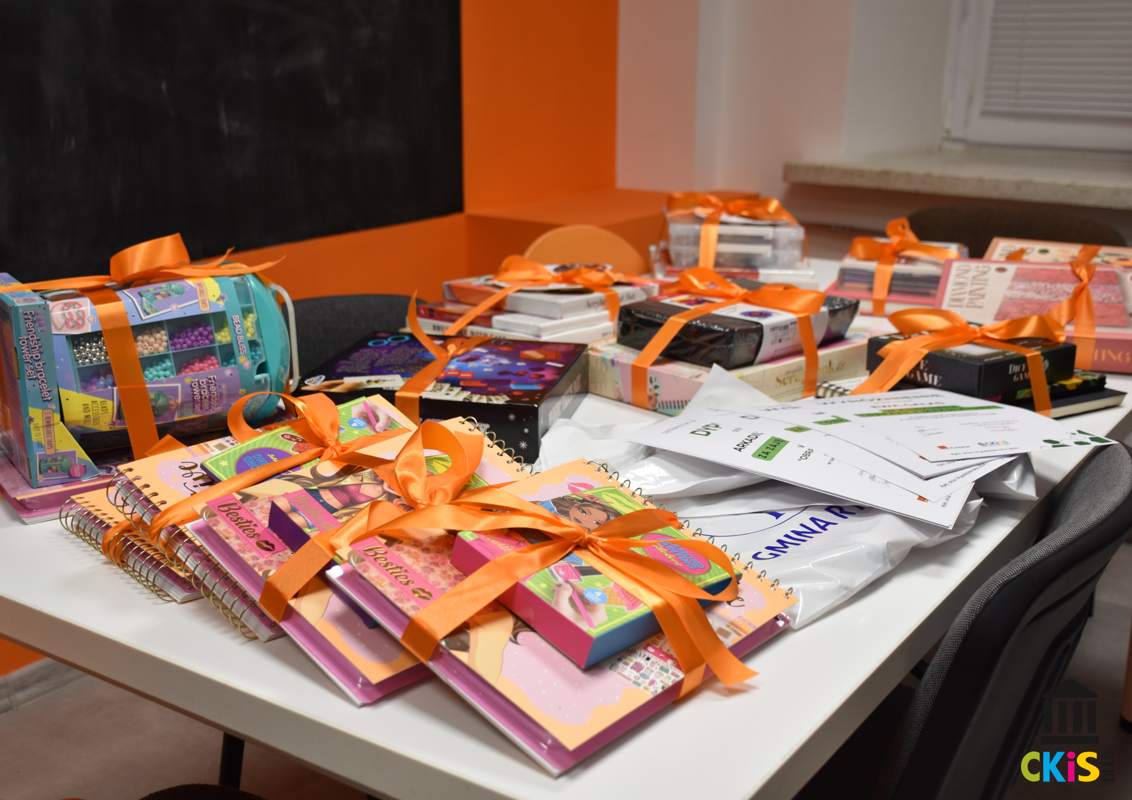Zdjęcie przedstawia stół w Pracowni Orange w Rykach cały zapełniony nagrodami dla laureatów konkursu. Każda paczka przewiązana jest pomarańczową wstążką. Na środki leżą dyplomy.