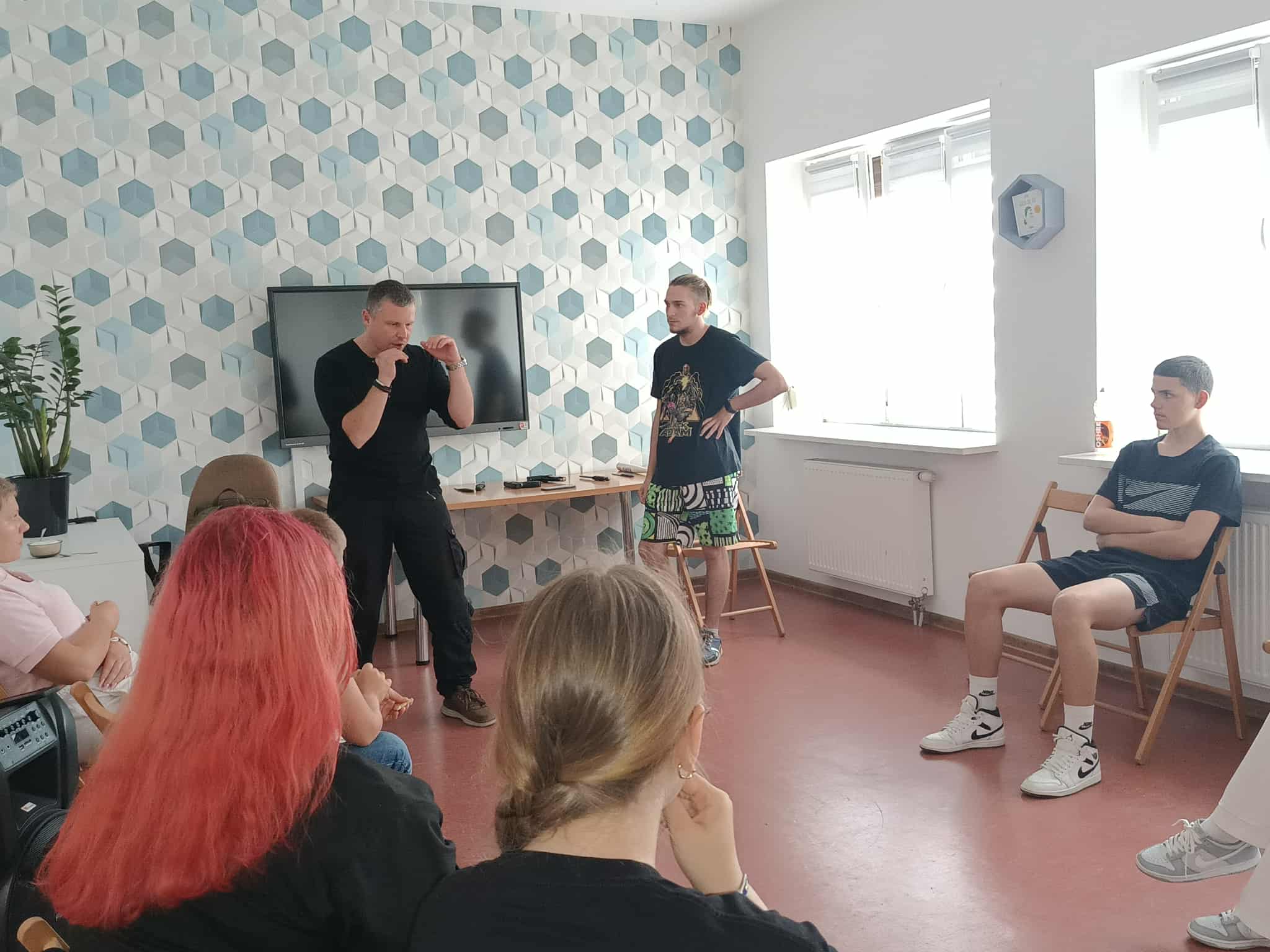 uczestnicy siedzą w kole na krzesełkach prowadzący z jednych chłopcem pokazuje podstawowe chwyty samoobrony