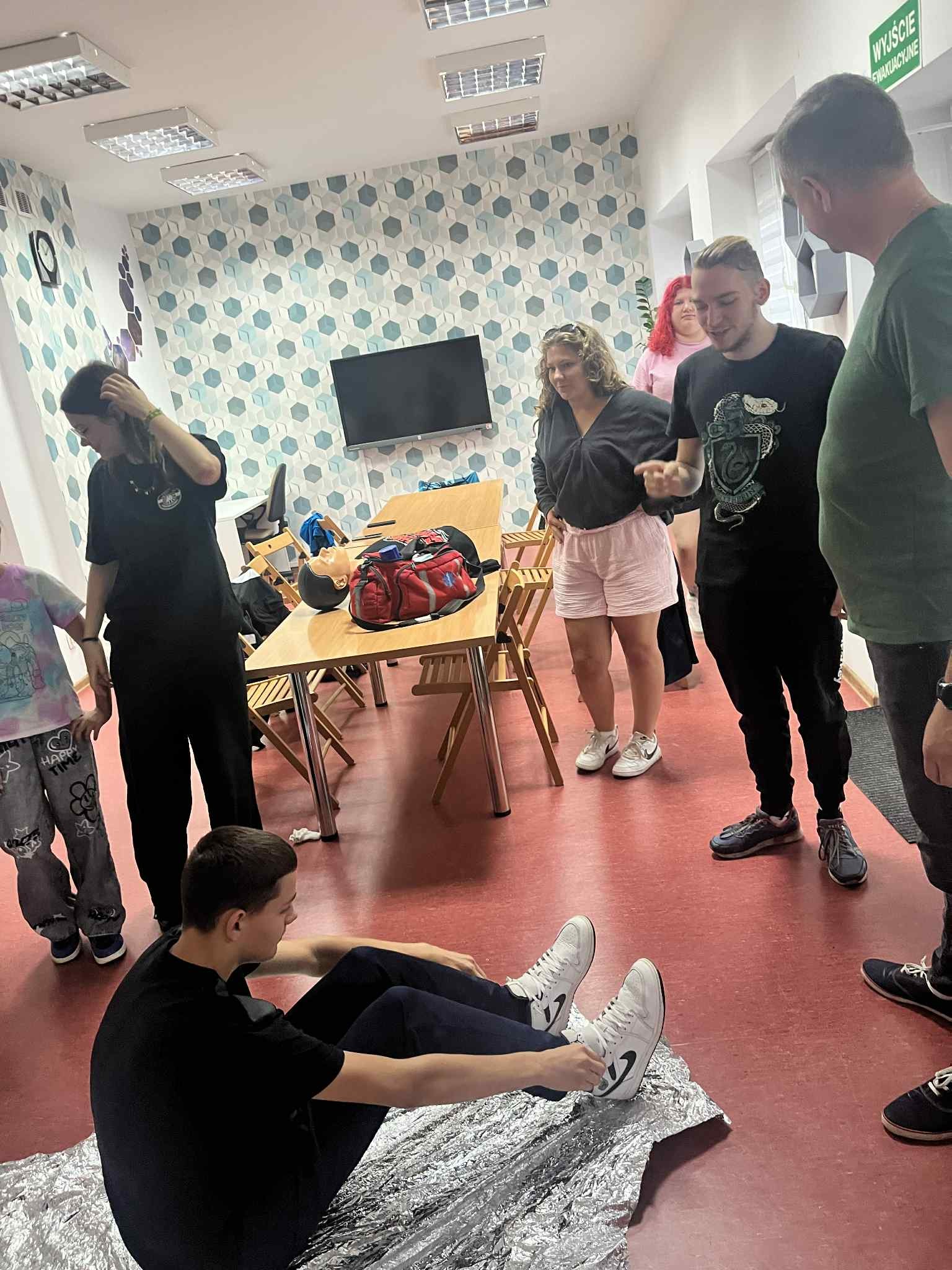 na podłodze leży folia życia a na niej siedzi uczestnik , pozostali obserwują