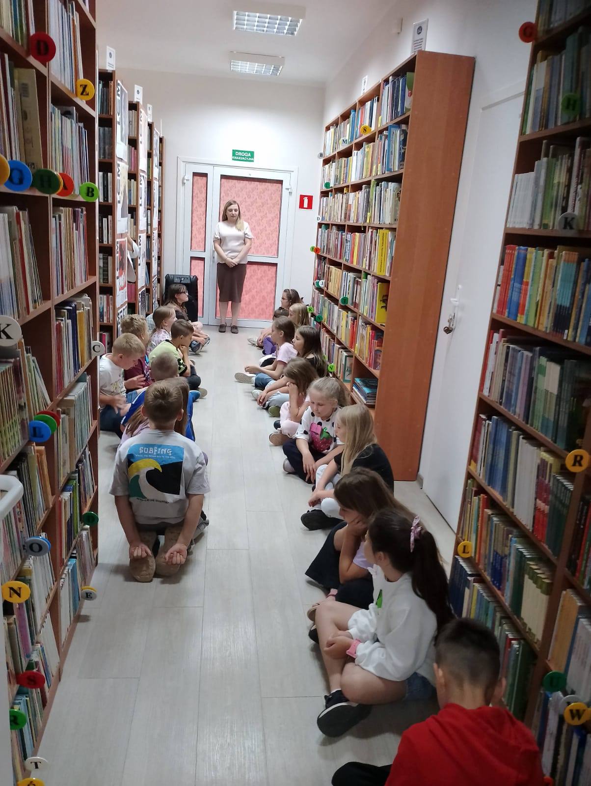 dzieci siedzą na holu bibliotecznym w 2 rzędach słuchając prowadzącej