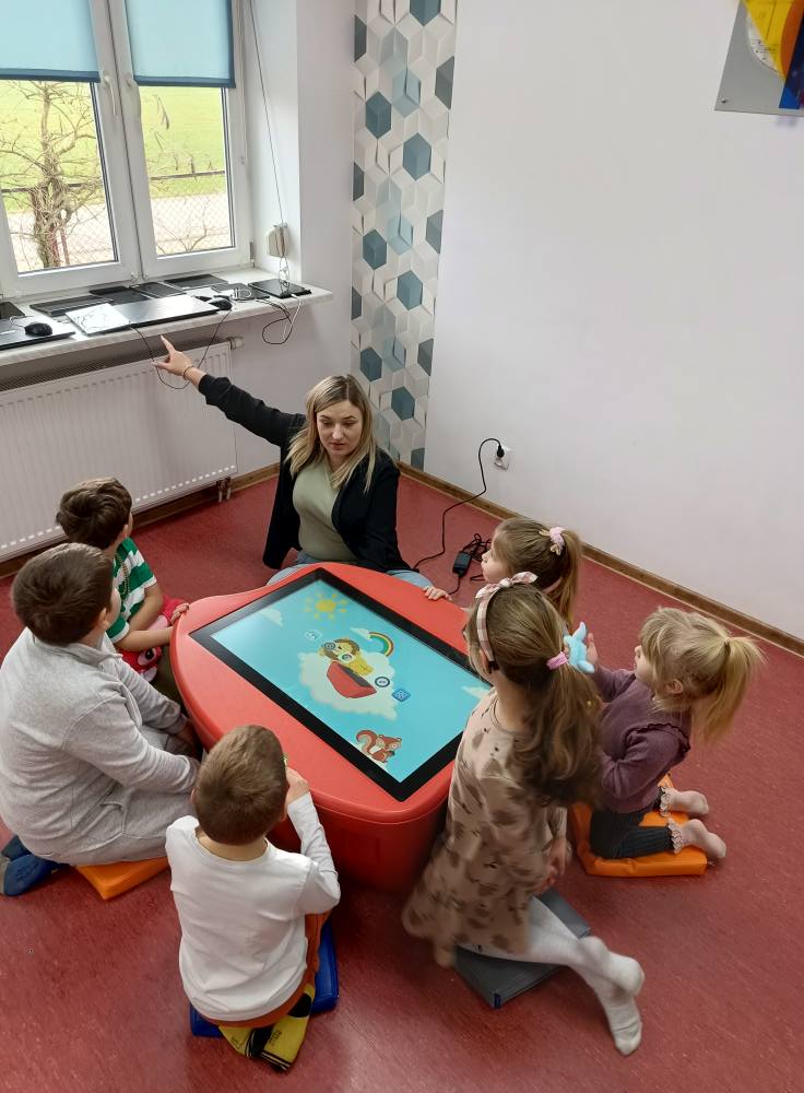 dzieci z prowadząca siedzą wokół stolika edukacyjnego