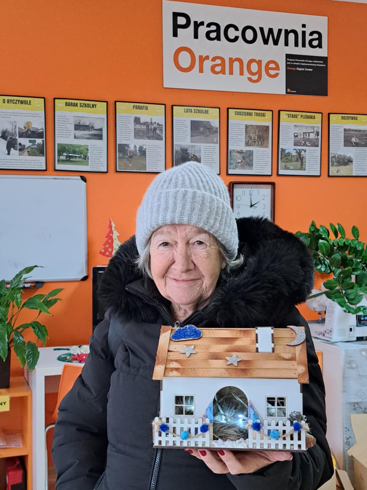 Pani trzyma w rękach drewniany domek, ozdobiony światecznie.