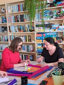 Dwie panie siedzą przy stole i pracują przy krepinie.