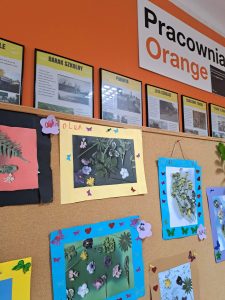 Na tle pomarańczowej ściany znajduje się wystawa prac dzieci - roślinne obrazy.