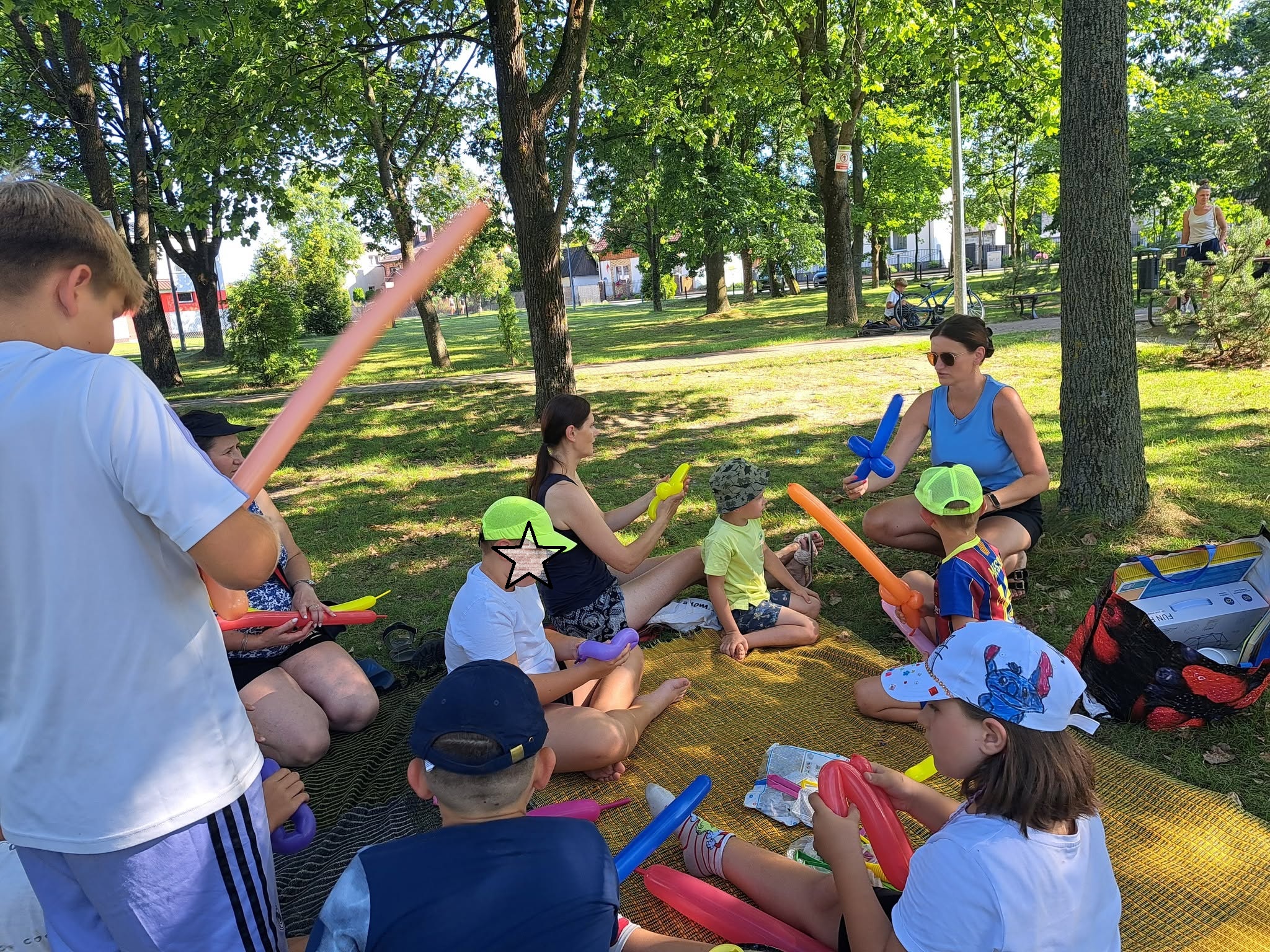 Na trawie siedzi grupa dzieci i dorosłych, modelują kolorowe balony.