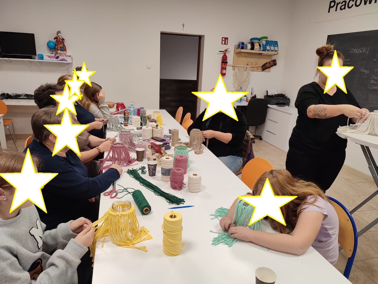 Grupa osób siedzi przy stole tworząc makramowe lampiony.