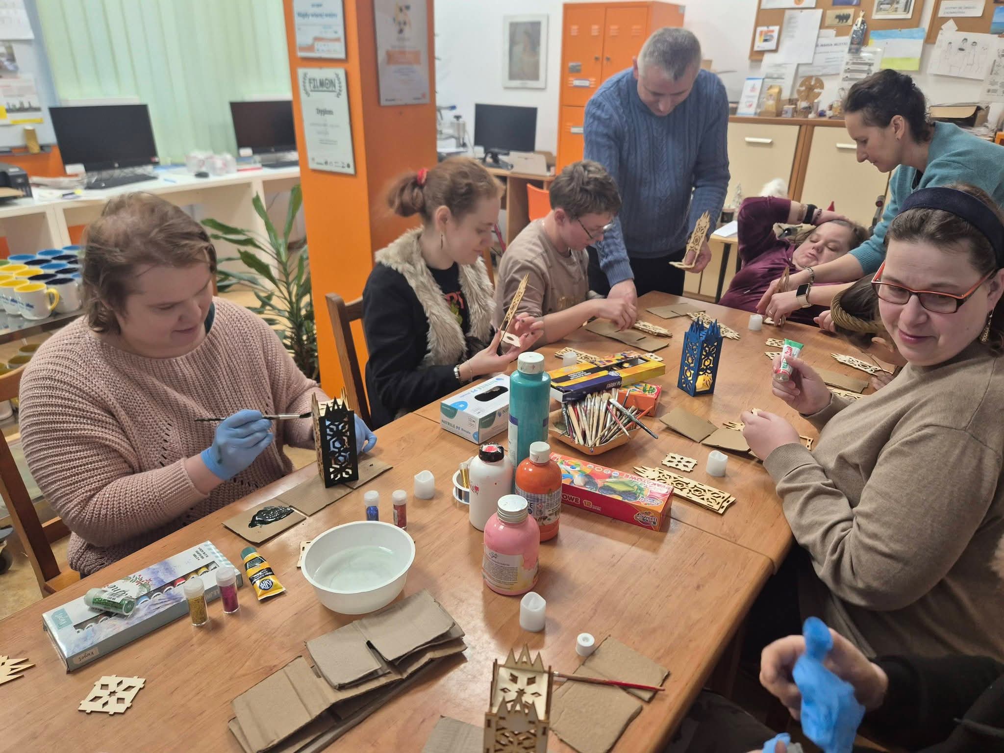 Grupa osób siedzi przy stole i maluje lampiony.