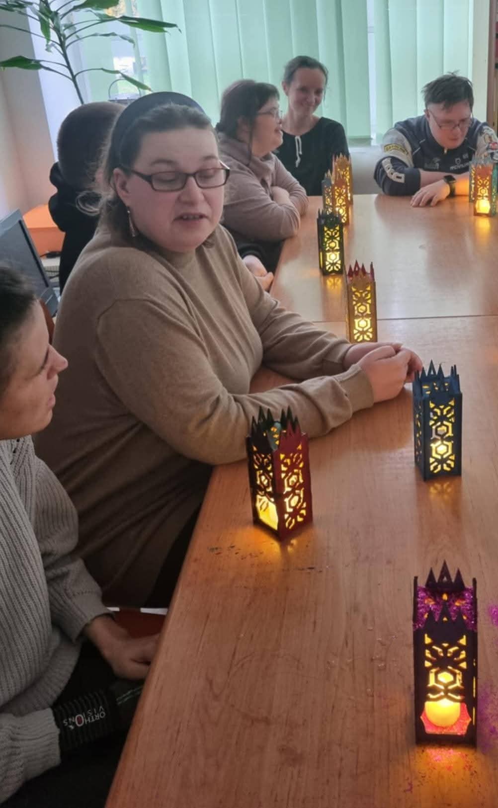 Grupa osób siedzi przy stole, obok nich stoją wykonane lampiony.