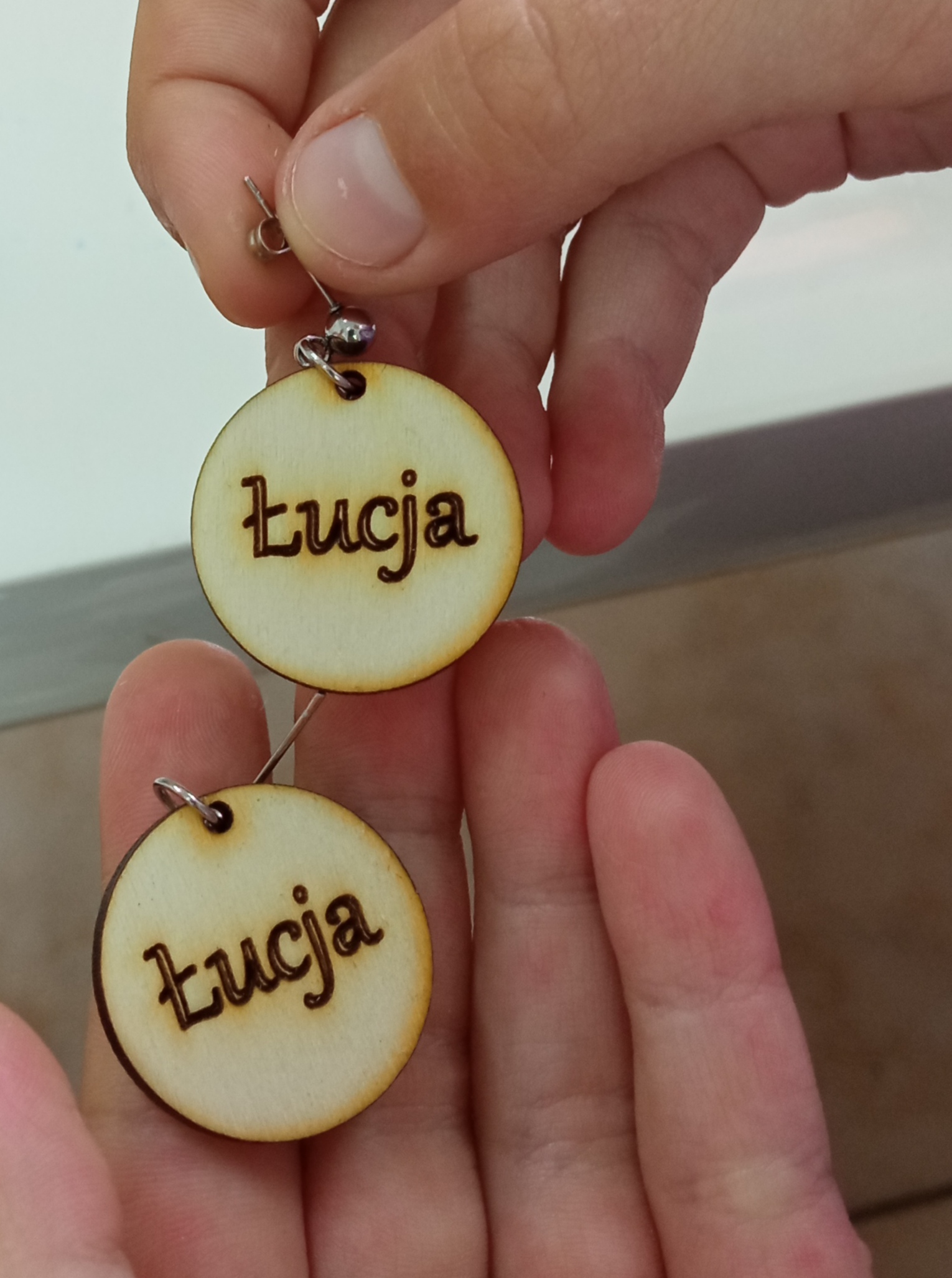 Okrągłe, drewniane kolczyki z napisem "Łucja" trzymane w dłoni.