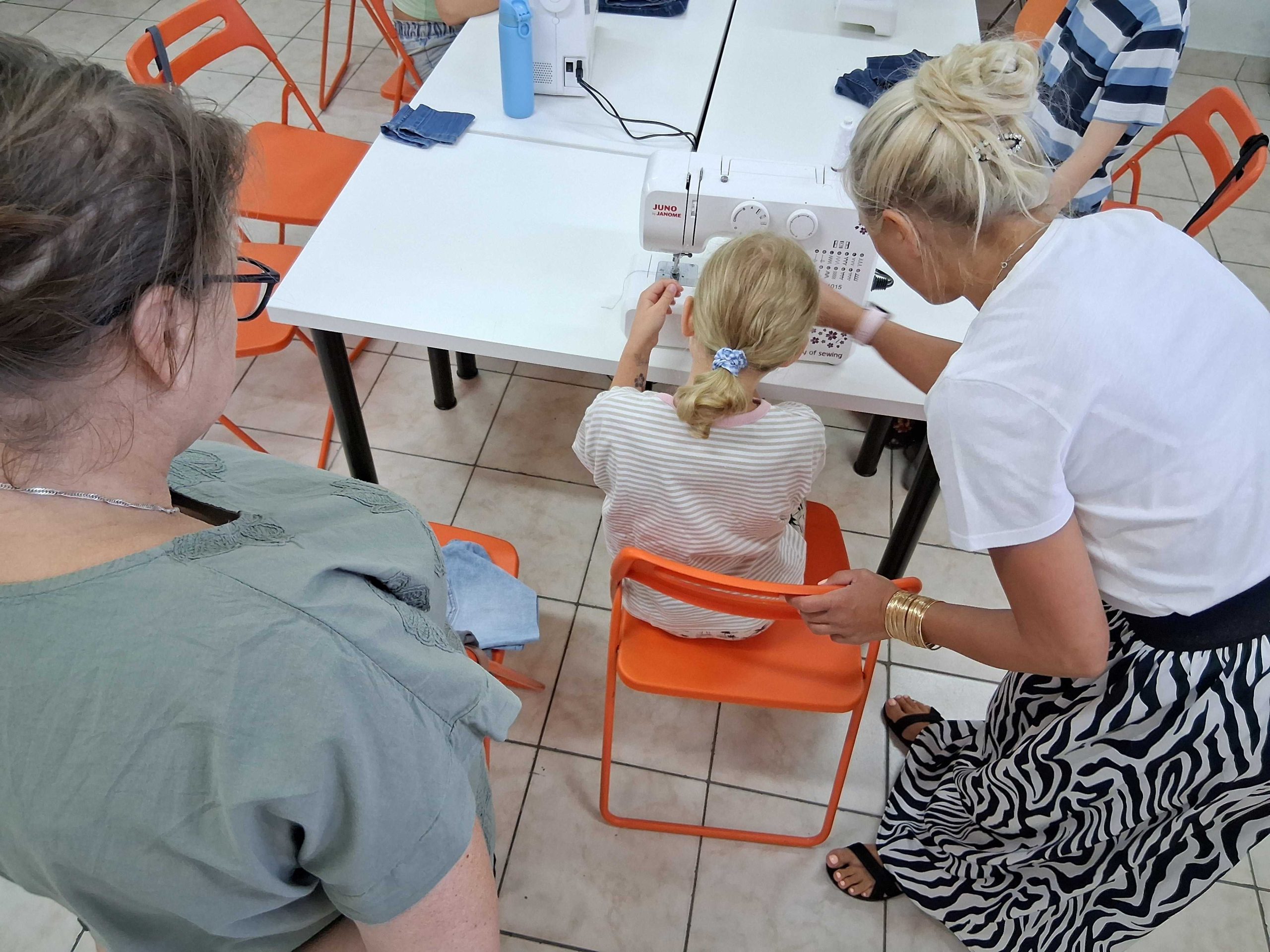 Na zdjęciu widać dziewczynkę siedzącą przy biurku z maszyną do szycia oraz instruktorkę, która coś jej tłumaczy