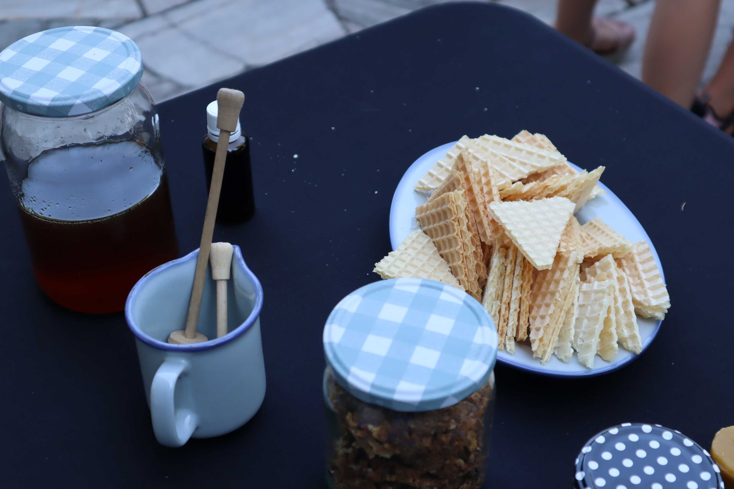 Zdjęcie przedstawia wafelki pokrojone w trójkąty, miód w słoiku i garnuszek z nabierakami do miodu leżące na czarnym stoliku
