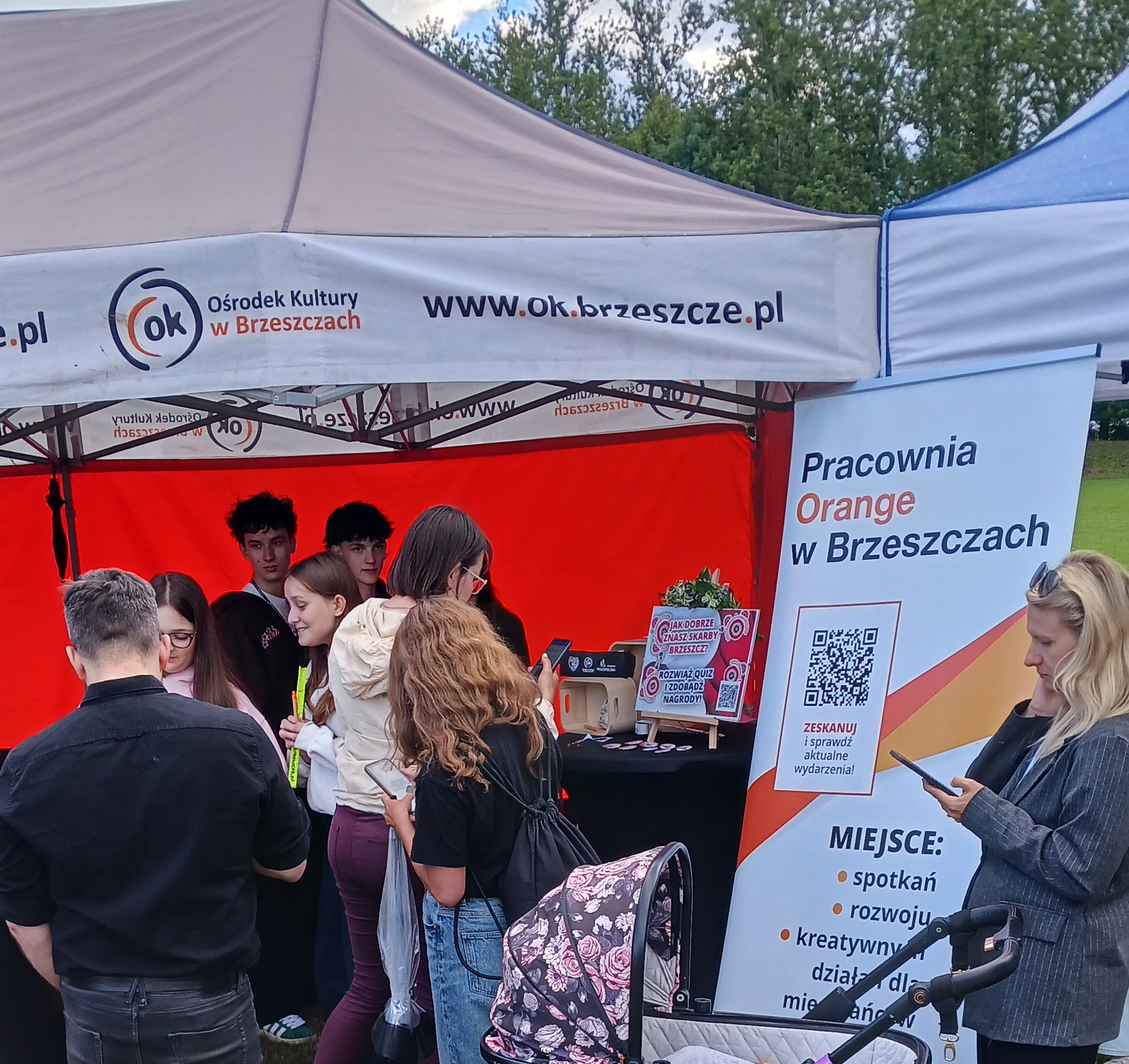 Na zdjęciu widać kilka osób skupionych przy stoisku plenerowym Pracowni Orange, pod namiotem, podczas wydarzenia Etno Skarby Brzeszcz.