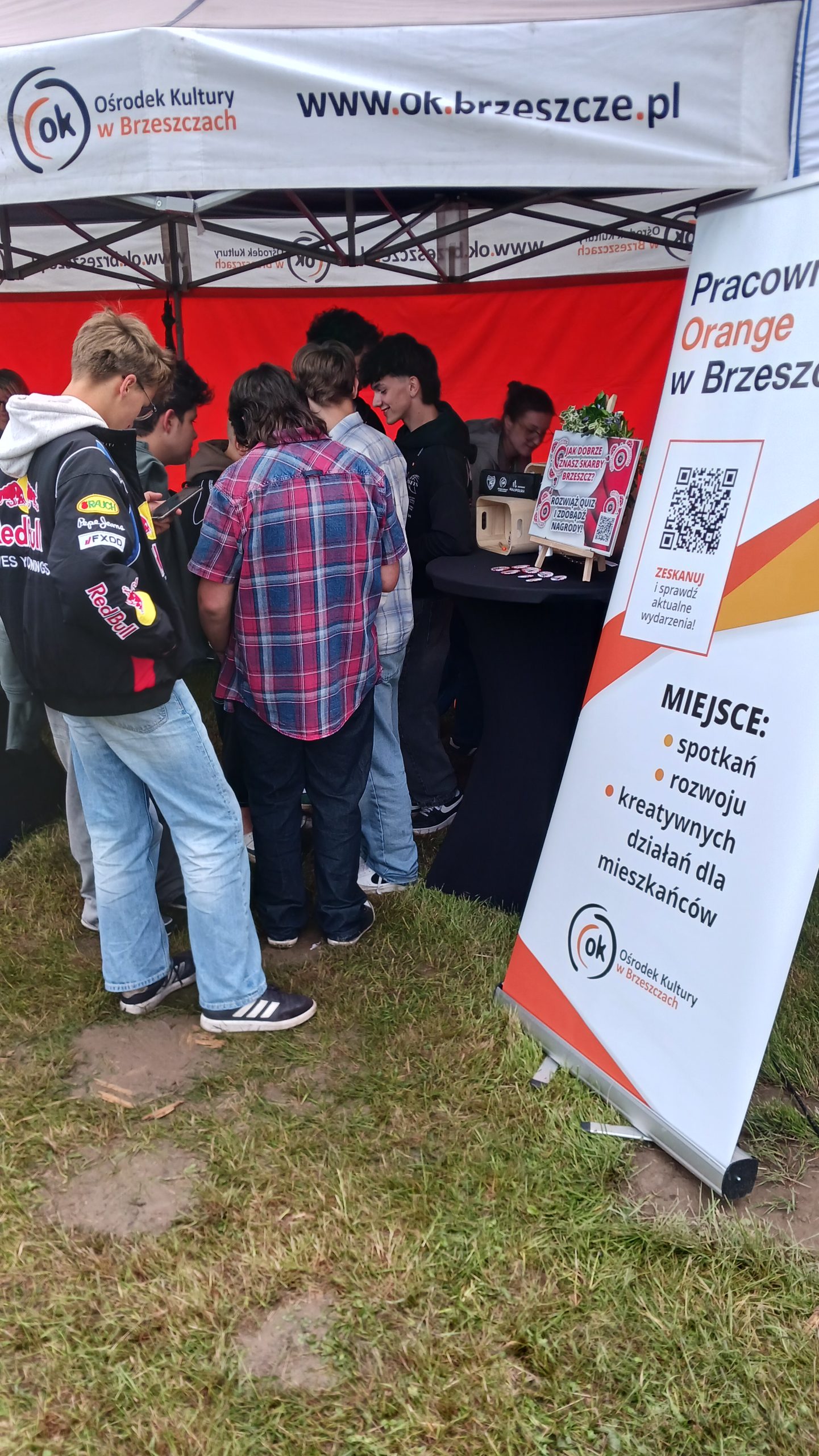 Widok zgromadzonej przy stoisku Pracowni Orange pod namiotem młodzieży. Po prawej stronie widoczny roll-up z napisem: Pracownia Orange w Brzeszczach, miejsce spotkań, rozwoju i kreatywnych działań dla mieszkańców