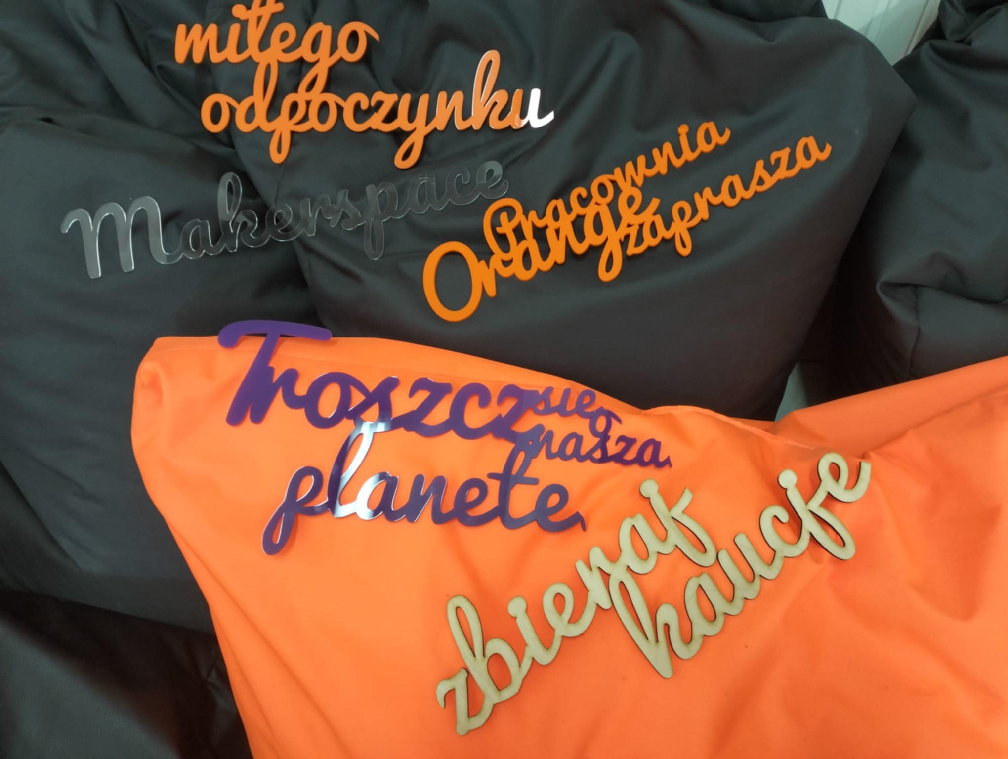 Napisy "miłego odpoczynku", "troszcz się o naszą planetę", "zbieraj kaucję", "makerspace", "Pracownia Orange zaprasza" wycięte z pleksi leżące na szarych i pomarańczowych pufach