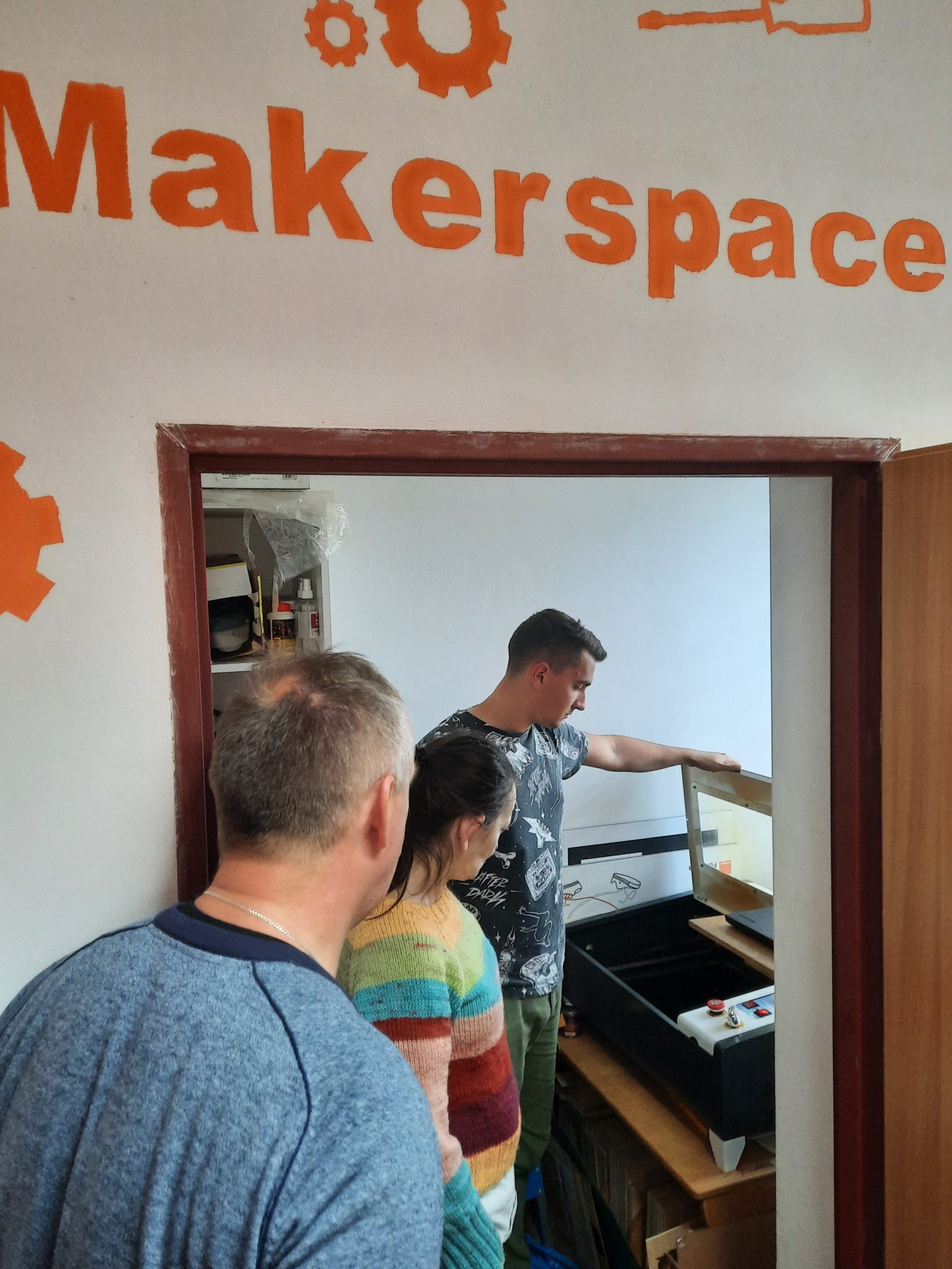 trzy osoby w pomieszczeniu podpisanym makerspace przygląda się ploterowi