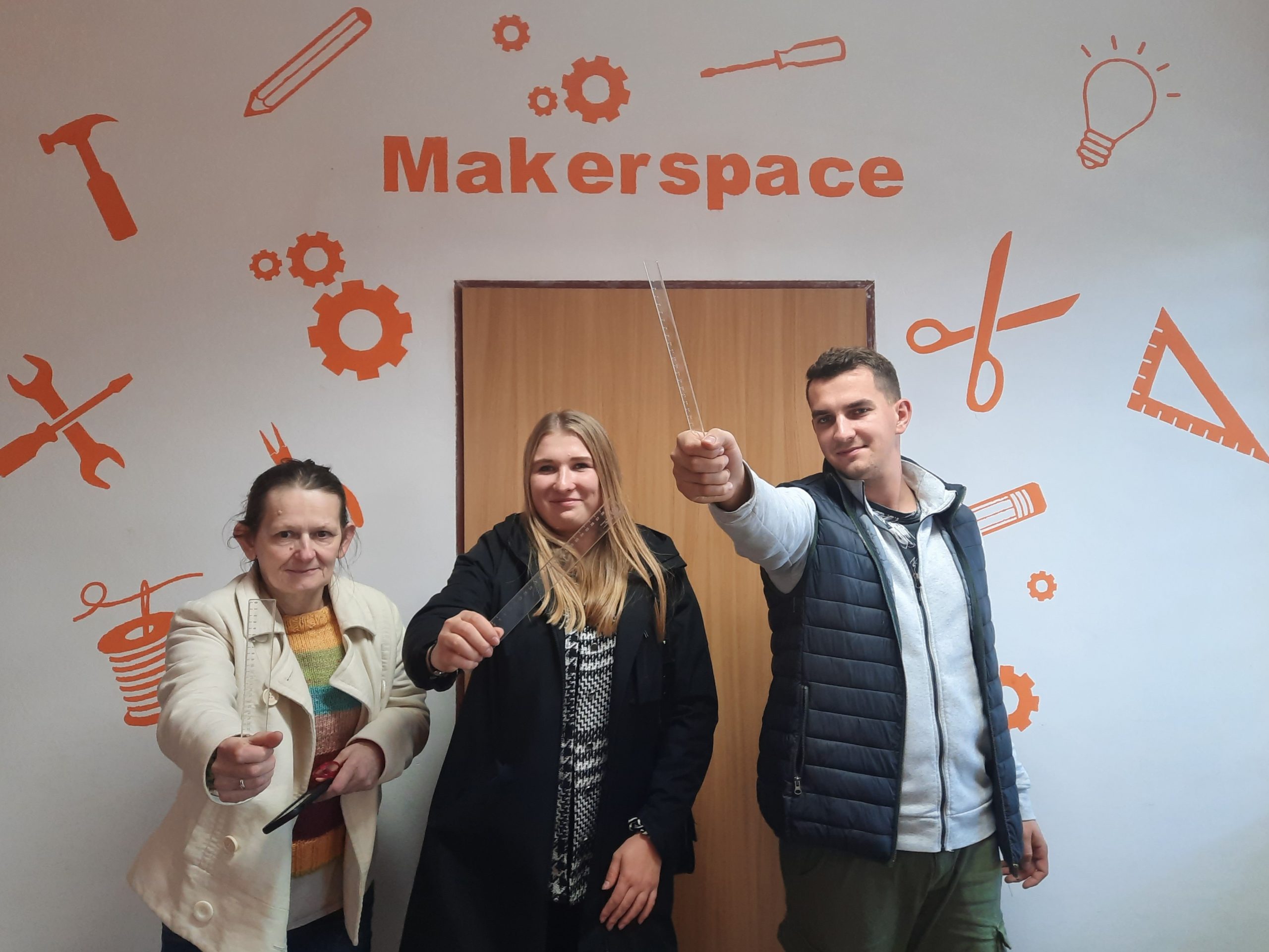 trzy osoby z wyciągniętymi linijkami na tle ściany z napisem makerspace