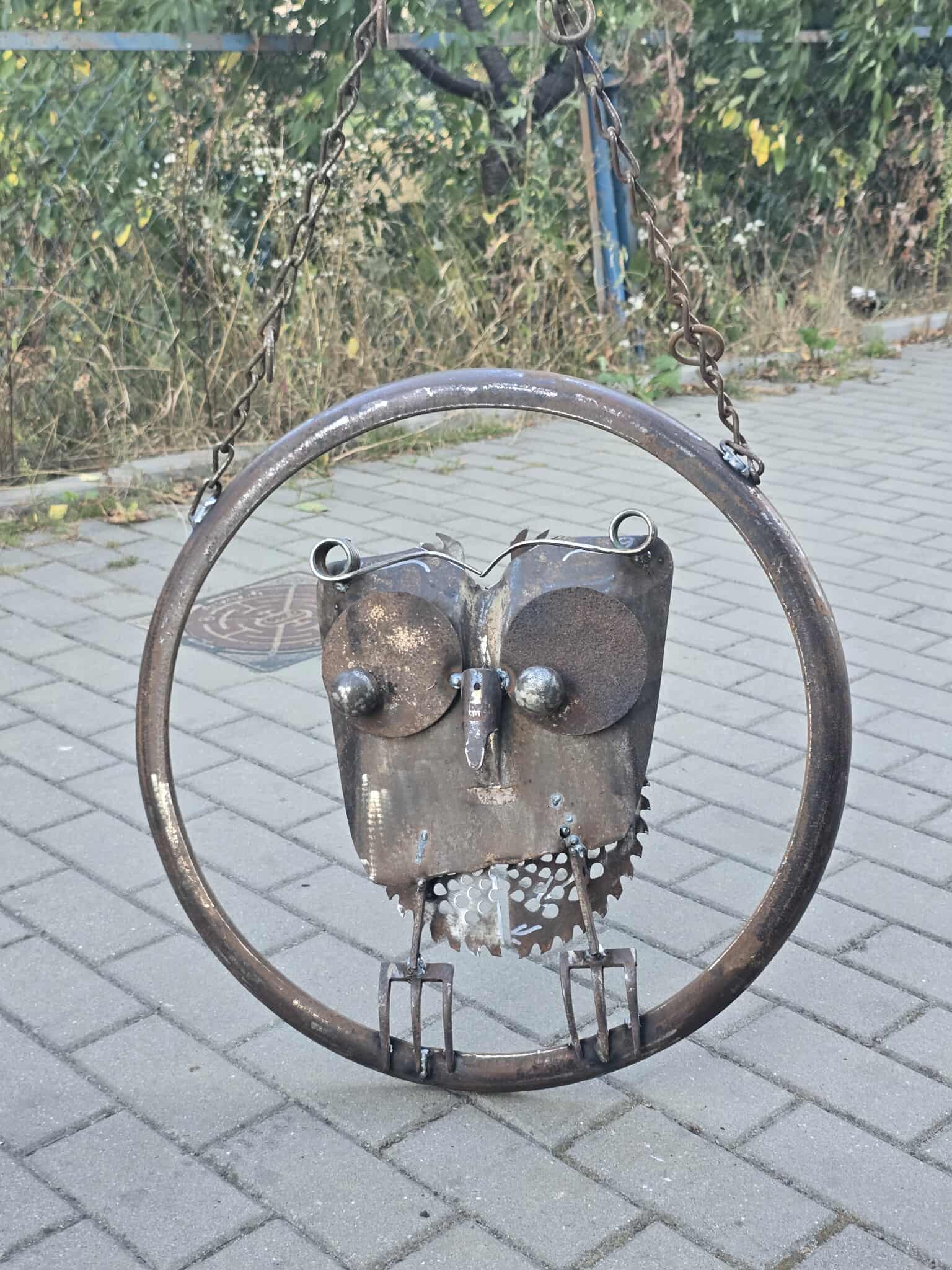 Metalowa rzeźba sowy wykonana z części mechanicznych, zawieszona na łańcuchach w okrągłej ramie. Konstrukcja stoi na tle chodnika z kostki brukowej i roślinności.