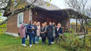 Grupa dziesięciu uśmiechniętych kobiet pozuje przed drewnianym domem w otoczeniu jesiennej przyrody. Kobiety stoją na trawie, ubrane w kurtki i szaliki, co sugeruje chłodną porę roku. W tle widoczny jest drewniany ganek oraz ozdobne elementy architektoniczne przy oknie i narożniku budynku.