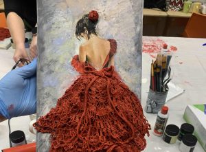 „Osoba trzyma obraz przedstawiający kobietę w czerwonej sukni z odkrytymi plecami. Dolna część sukni jest trójwymiarowa i wykonana z koronkowej, czerwonej tkaniny. W tle widać stół warsztatowy z farbami, pędzlami i narzędziami, a na rękach osoby trzymającej obraz znajdują się rękawiczki ochronne.”