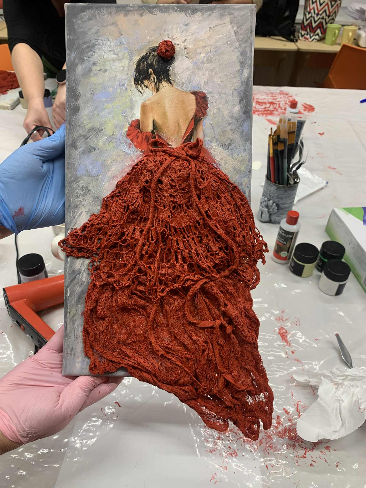 Osoba trzyma obraz przedstawiający kobietę w czerwonej sukni z odkrytymi plecami. Dolna część sukni jest trójwymiarowa i wykonana z koronkowej, czerwonej tkaniny. W tle widać stół warsztatowy z farbami, pędzlami i narzędziami, a na rękach osoby trzymającej obraz znajdują się rękawiczki ochronne.