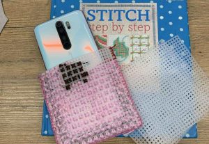 Na drewnianym blacie leży książka „Stitch Step by Step” w niebieskiej okładce w białe kropki. Na niej znajduje się smartfon w jasnej obudowie częściowo wsunięty w różowy, plastikowy, kanwasowy pokrowiec do haftu krzyżykowego z zaczętą różowo-brązową robótką. Obok leżą dwie plastikowe siatki kanwy do haftu — jedna różowa, druga przezroczysta.