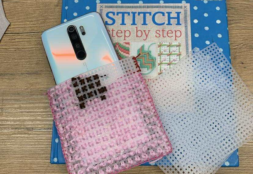 Na drewnianym blacie leży książka „Stitch Step by Step” w niebieskiej okładce w białe kropki. Na niej znajduje się smartfon w jasnej obudowie częściowo wsunięty w różowy, plastikowy, kanwasowy pokrowiec do haftu krzyżykowego z zaczętą różowo-brązową robótką. Obok leżą dwie plastikowe siatki kanwy do haftu — jedna różowa, druga przezroczysta.